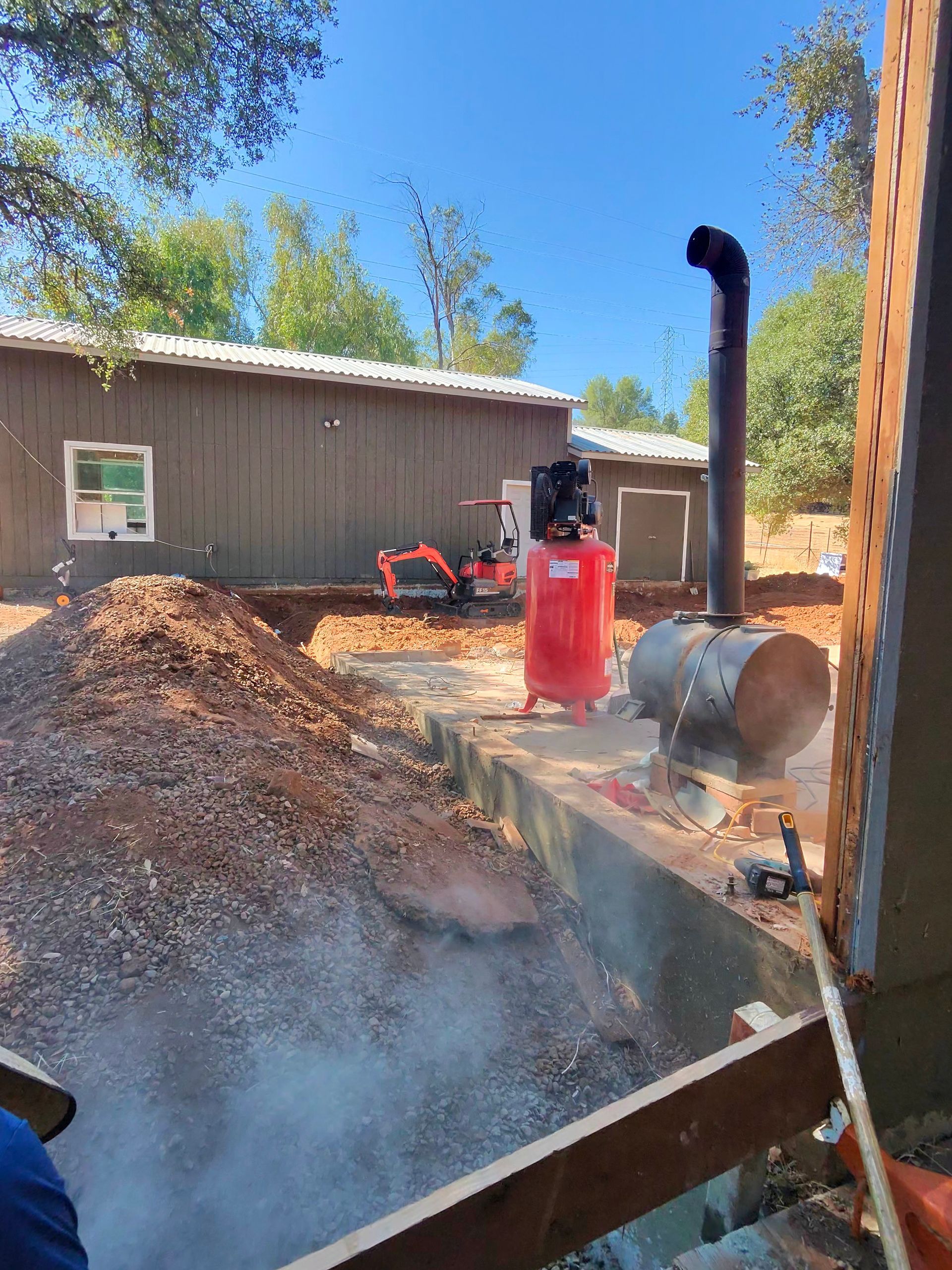 Outdoor wood chipper blowing smoke beside a shed, with a red tank and piles of mulch in a yard.