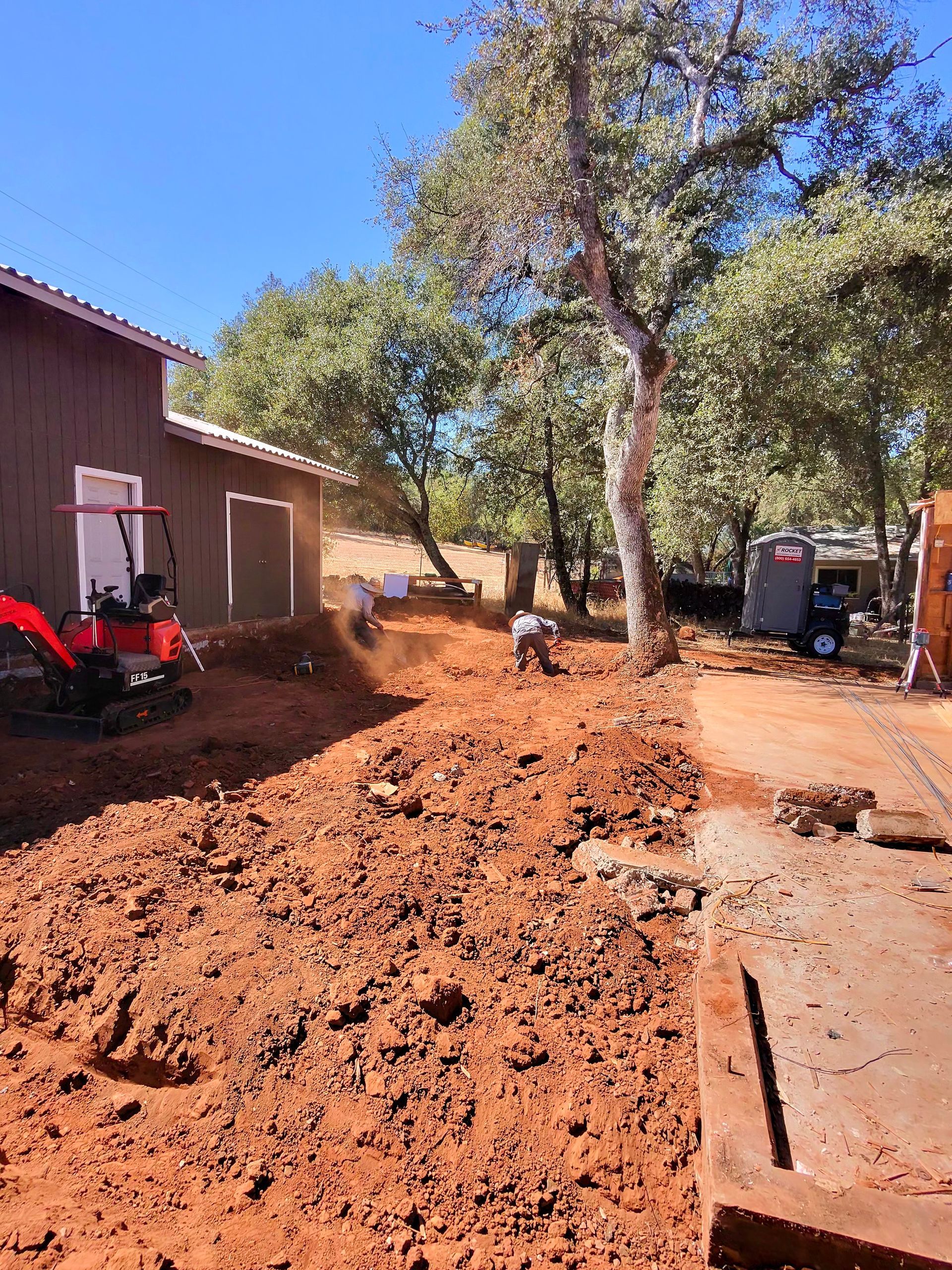Construction site with a red skid steer moving dirt beside a building and trees.