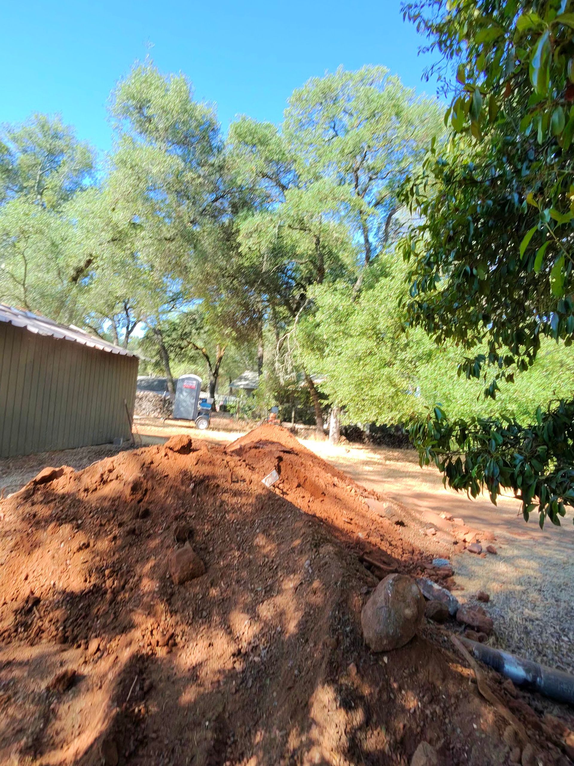 Pile of reddish dirt beside a building and trees under a bright blue sky