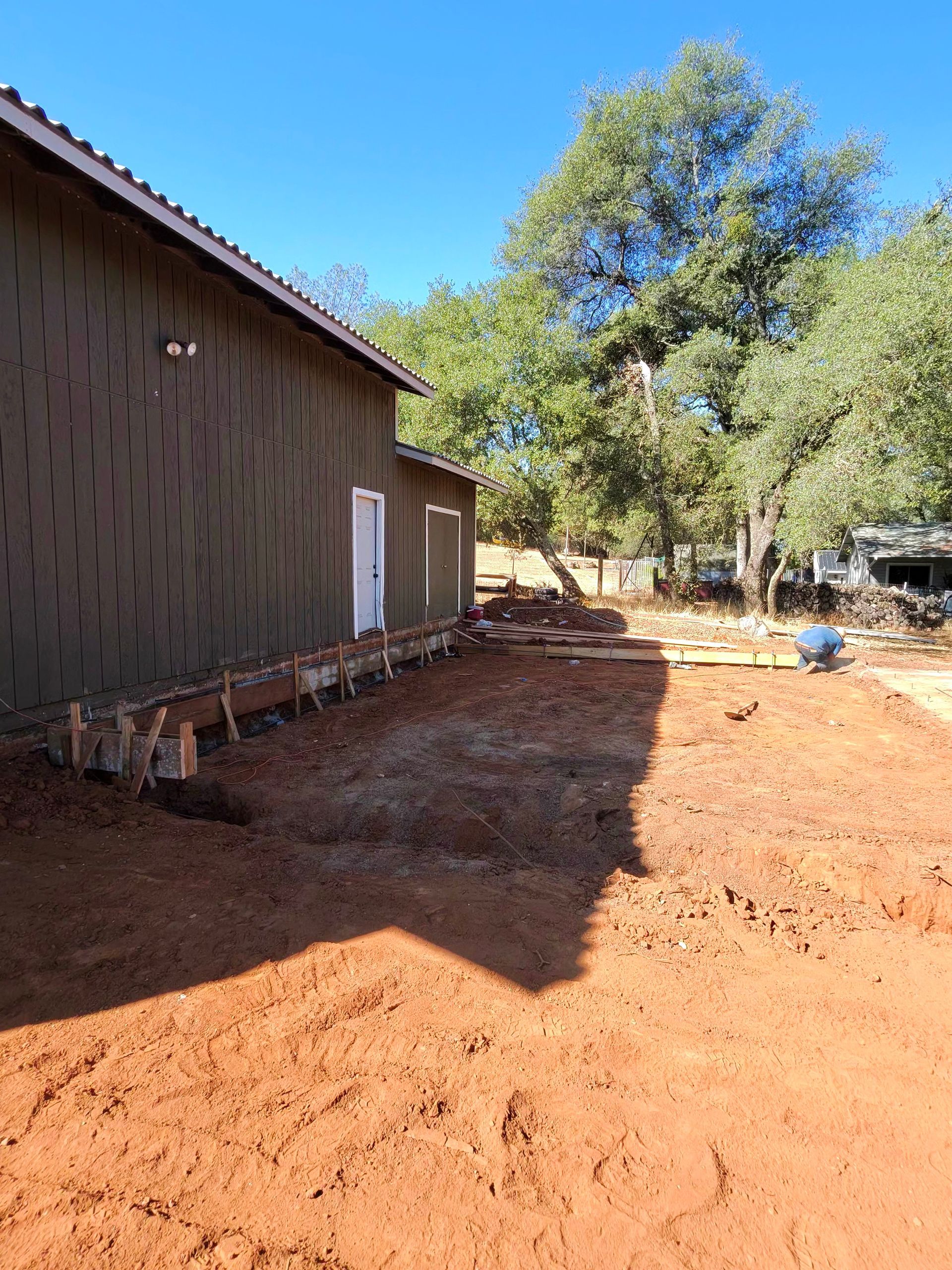 Rural yard beside a brown building, with red dirt ground and trees under a clear blue sky