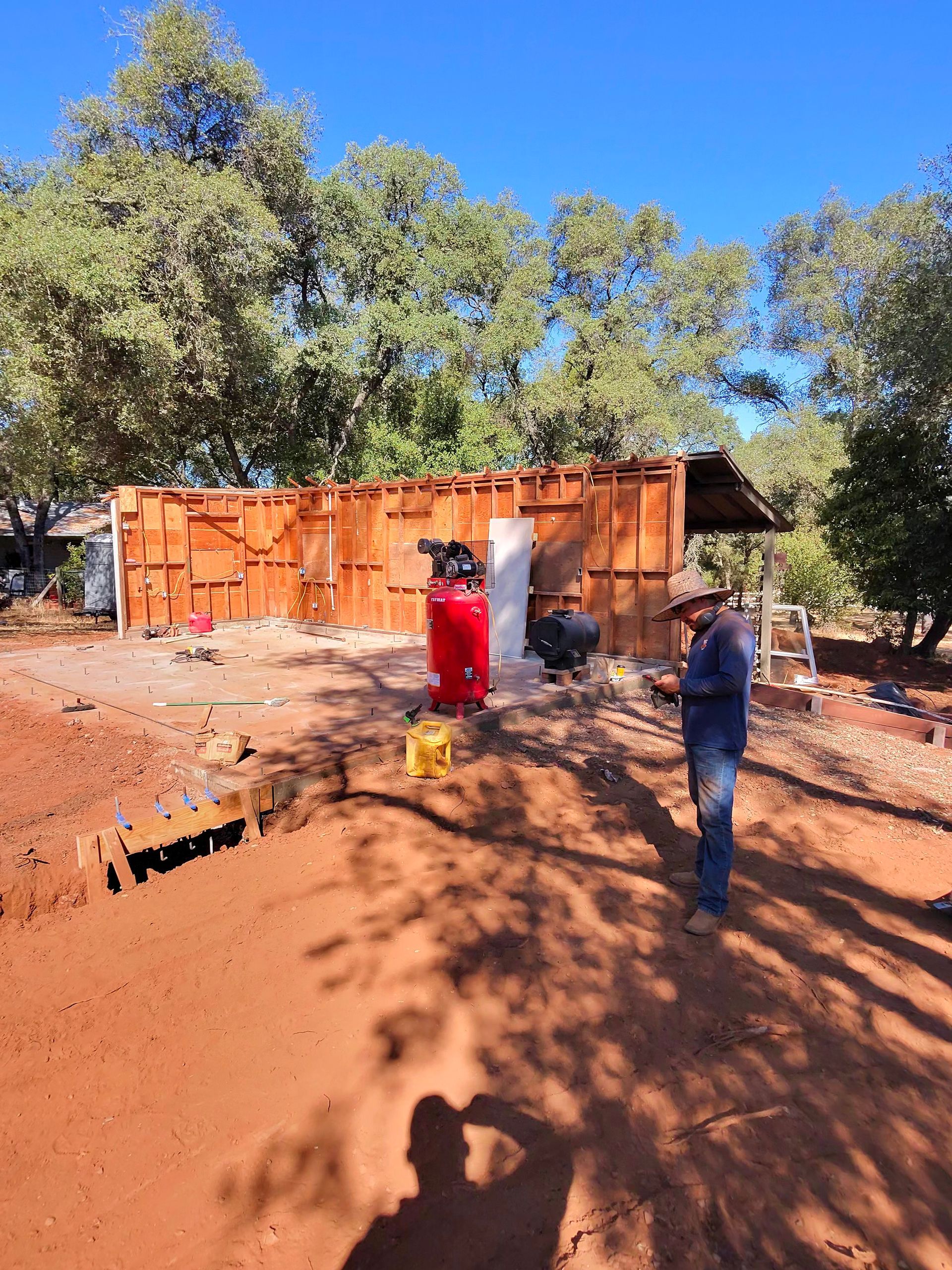 Two people at a wooden construction site, one spraying water near a red barrel under trees.