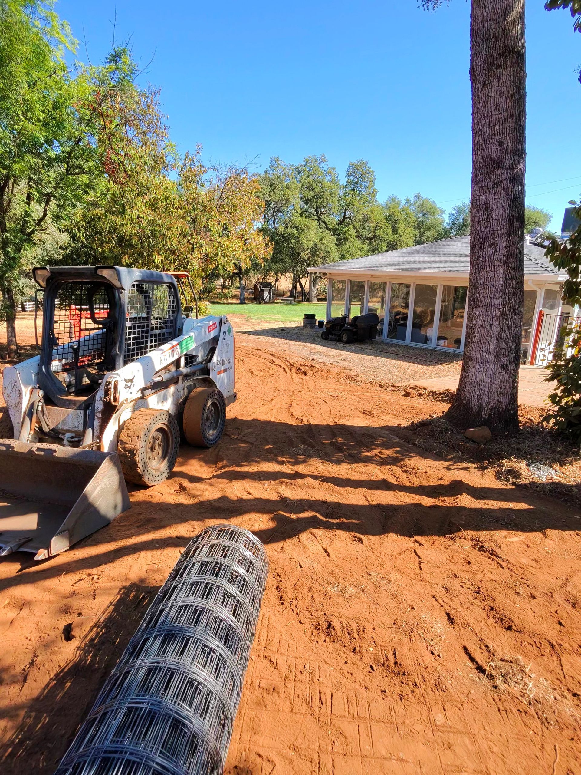 Skid steer leveling red dirt beside a house and trees on a sunny day