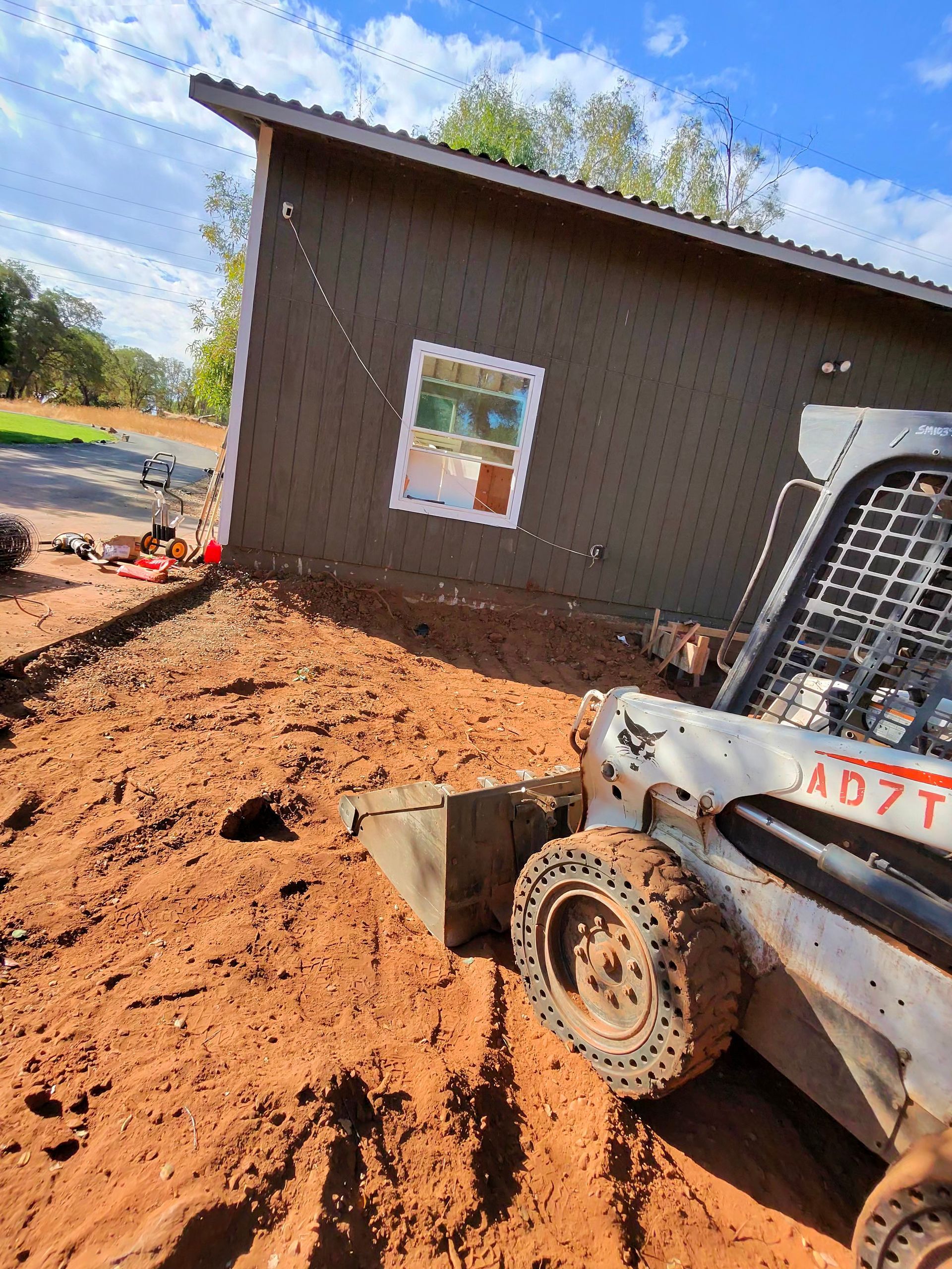 Skid steer pushing red dirt beside a brown house under construction with a window and sloped roof