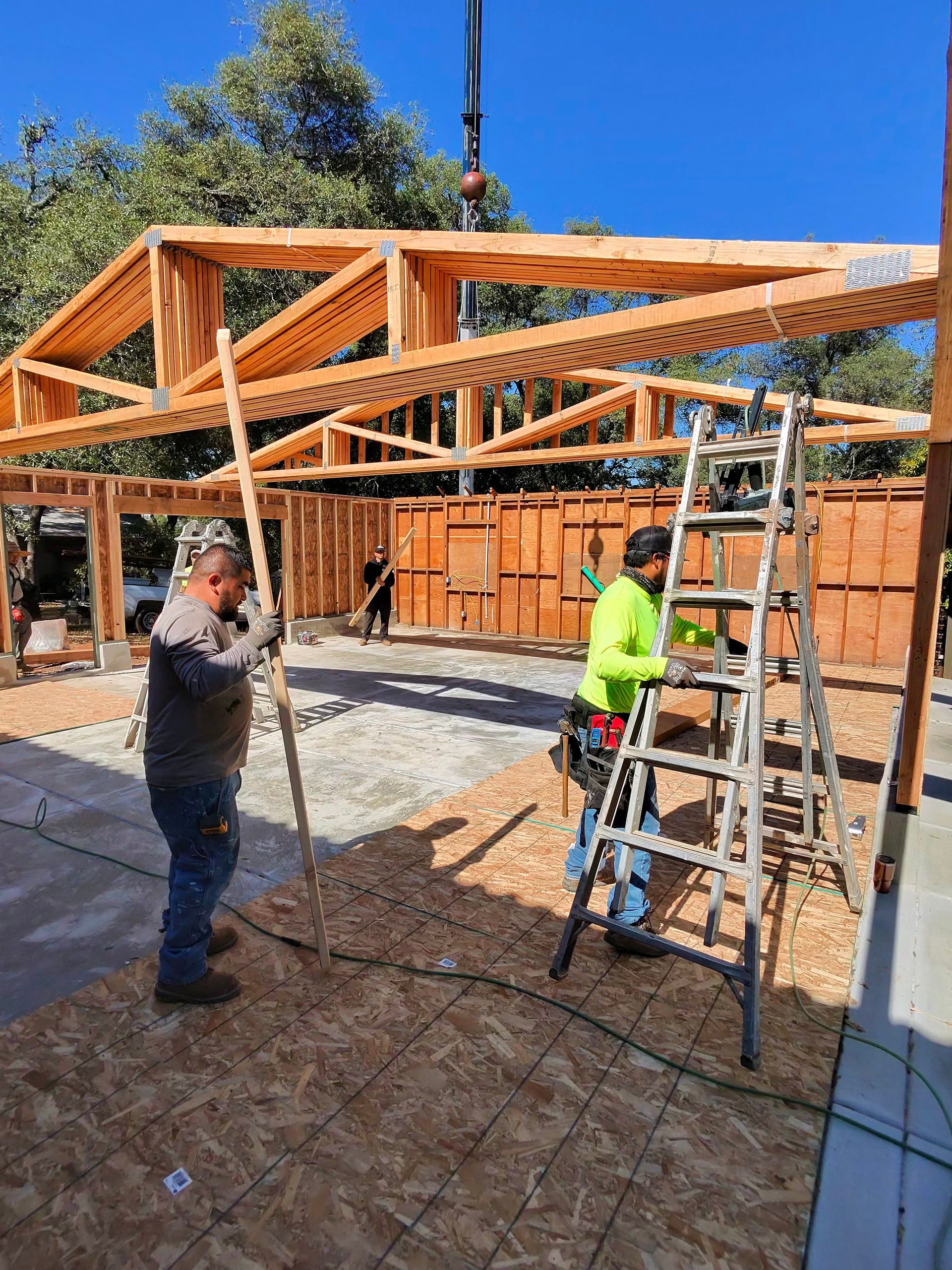 Workers assembling a wooden roof frame on a construction site, with ladders and scaffolding under a blue sky