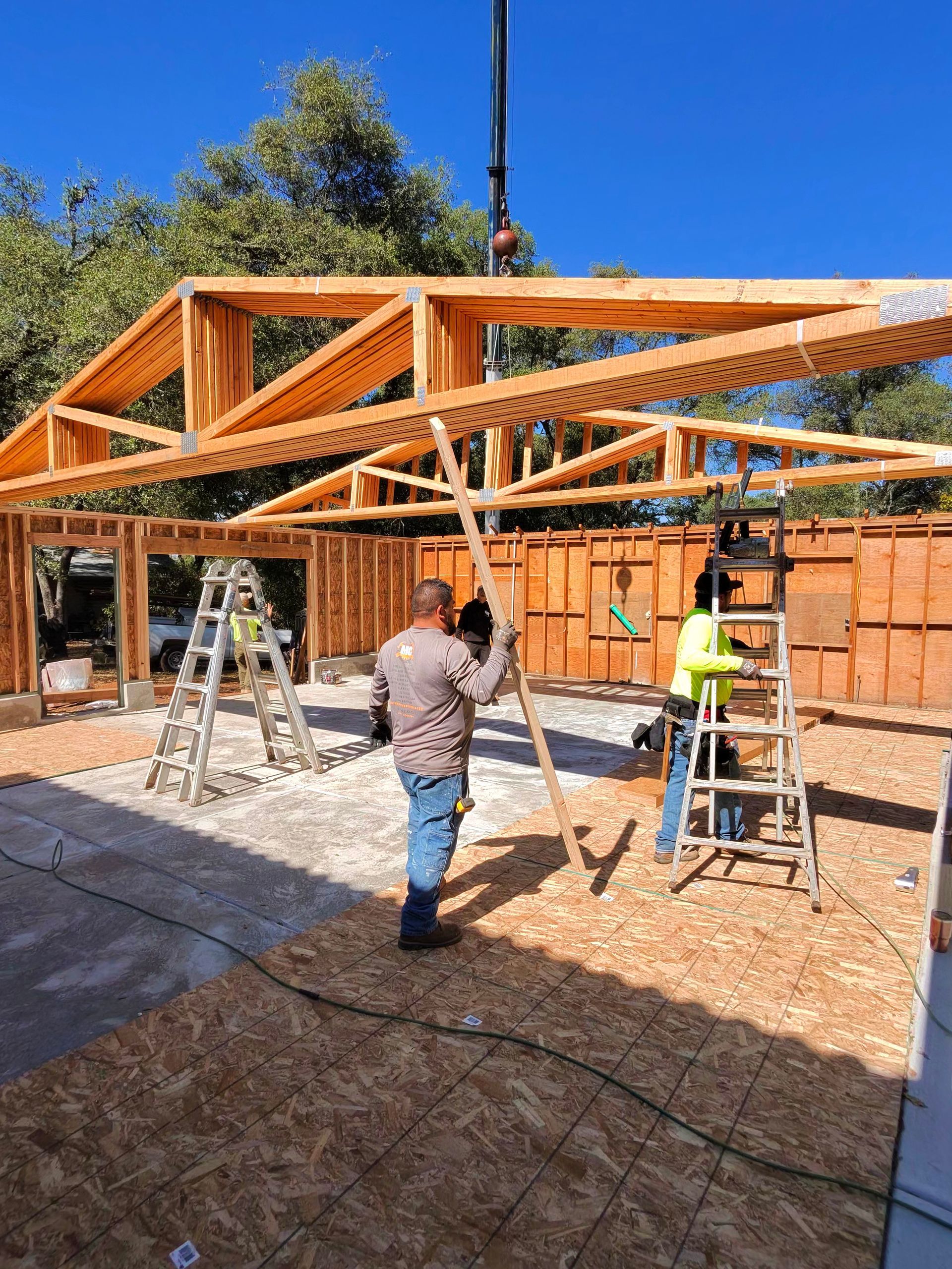 Workers assembling a large wooden roof frame beside a brick house on a sunny day