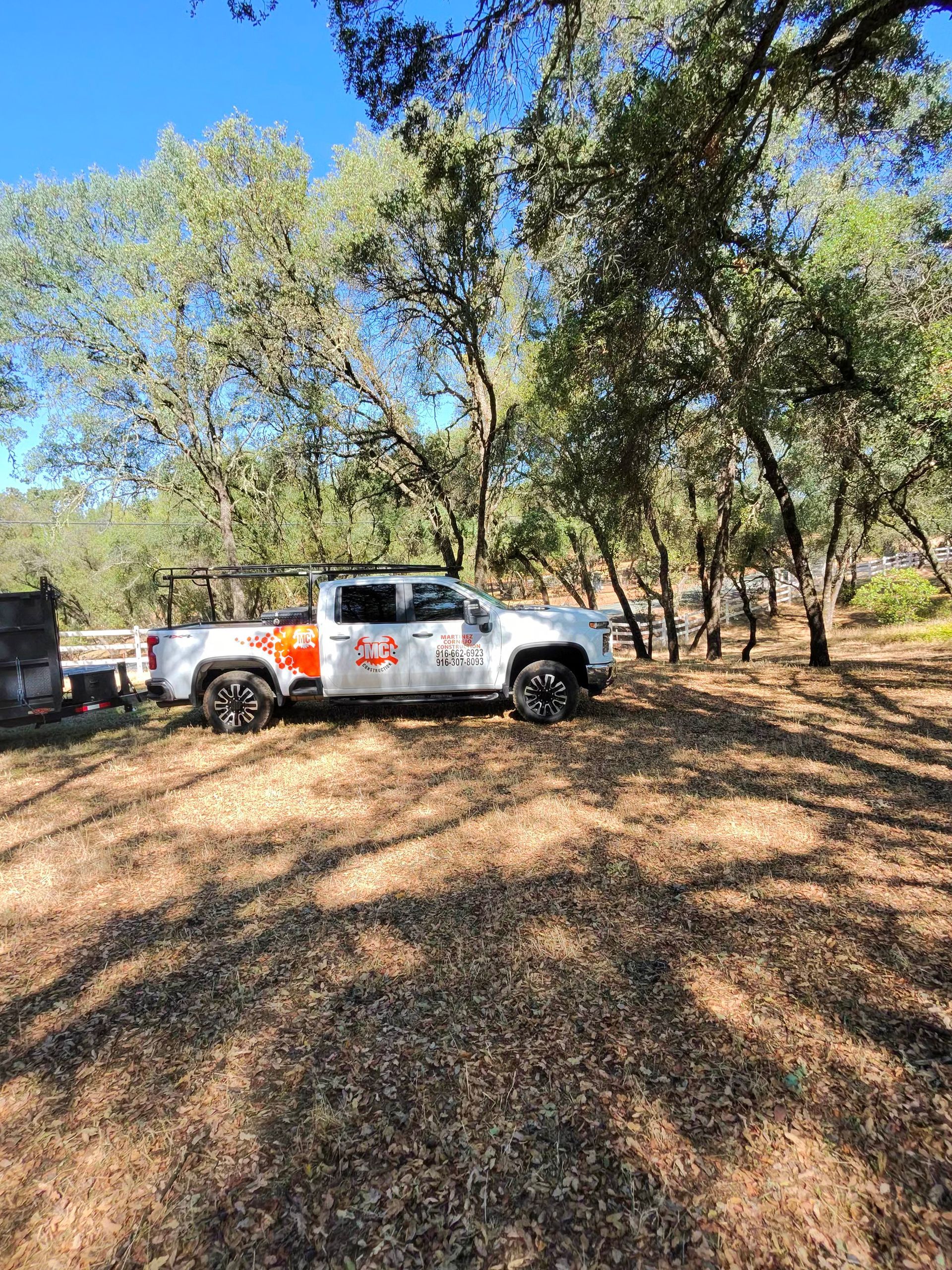 White pickup truck parked under trees on a leaf-covered path in a sunny wooded area.