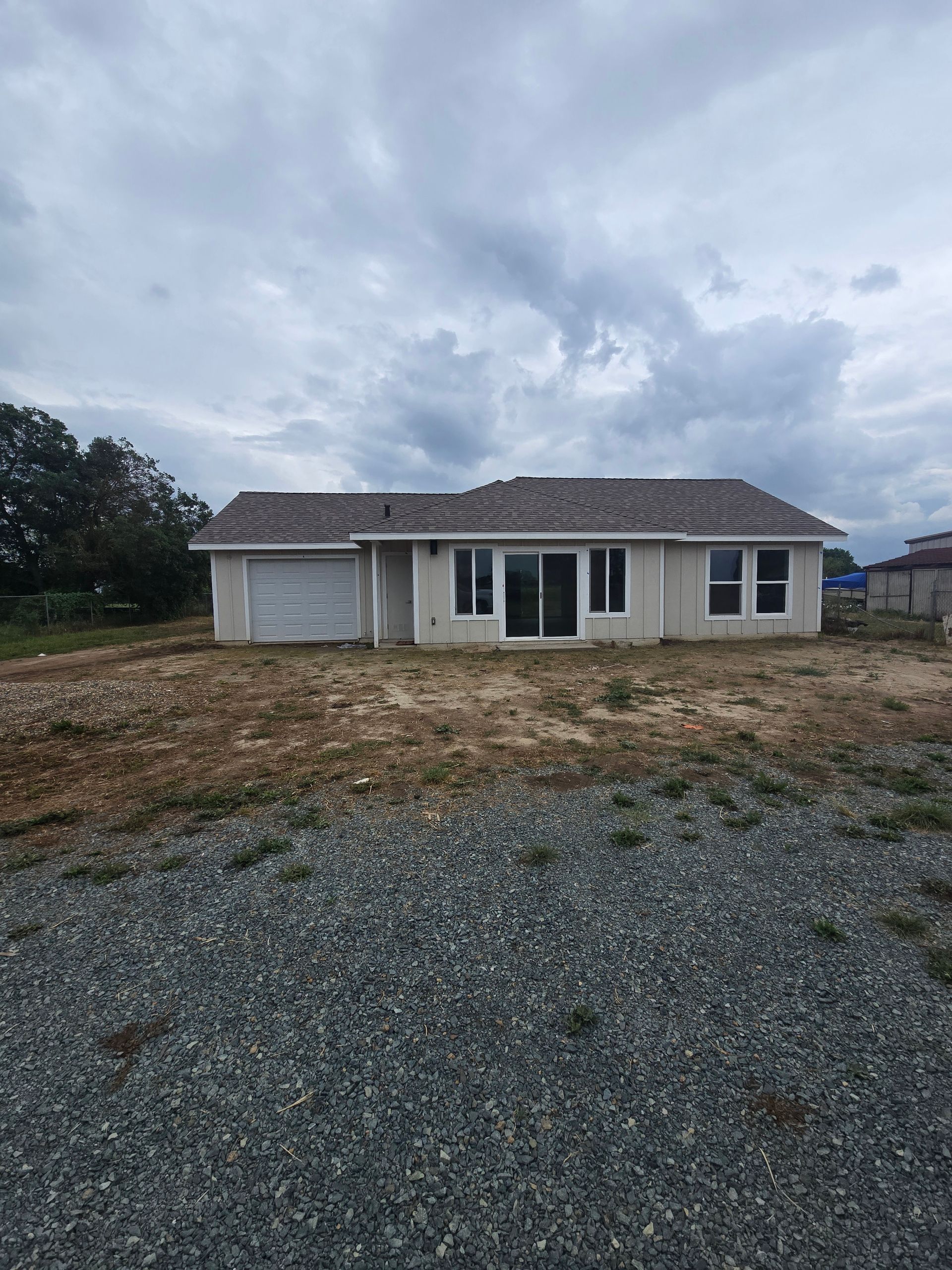 Single-story beige house with a garage and gravel driveway under a cloudy sky.