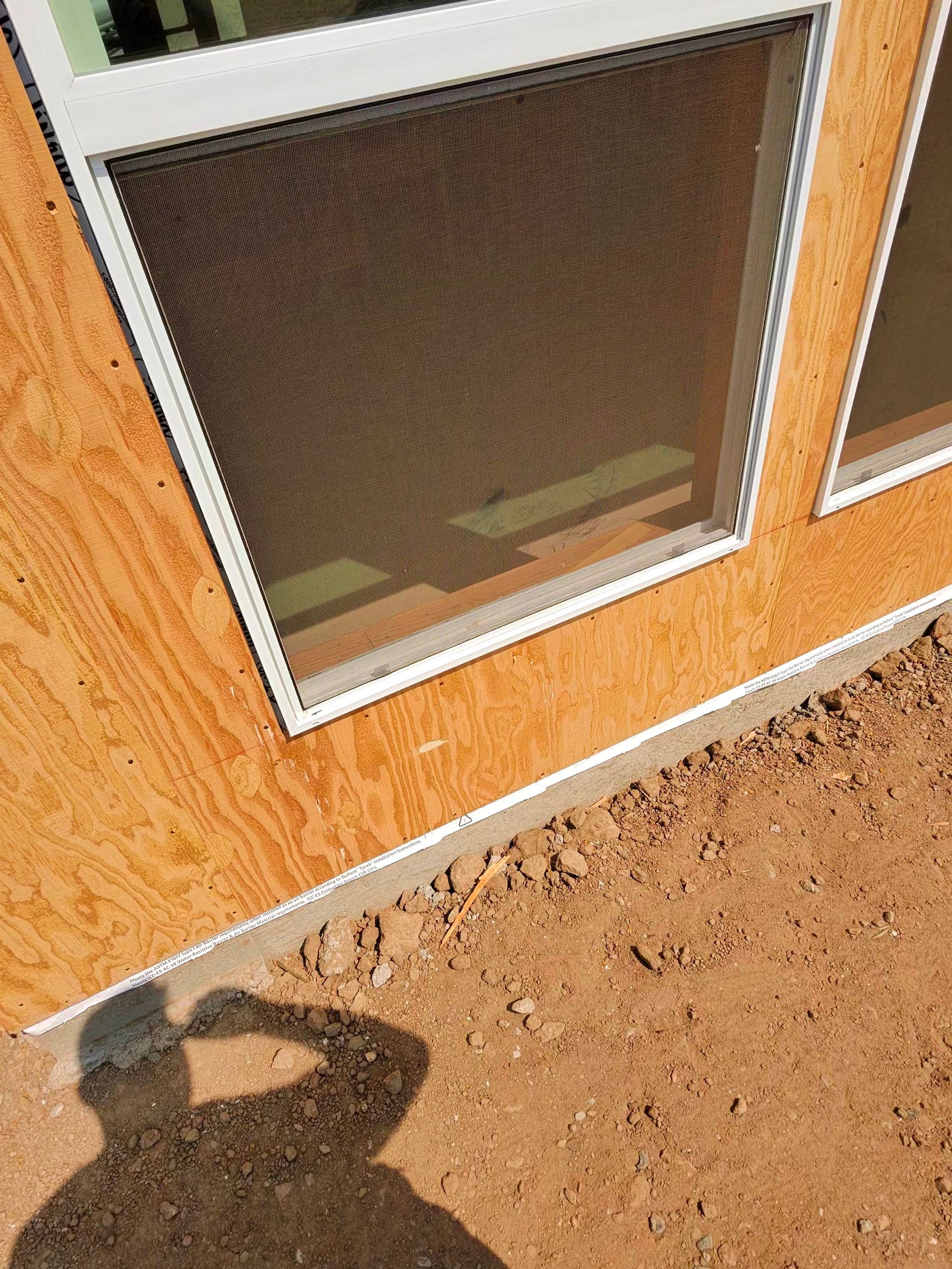 Orange stucco wall with a screened window above dirt ground and a person’s shadow