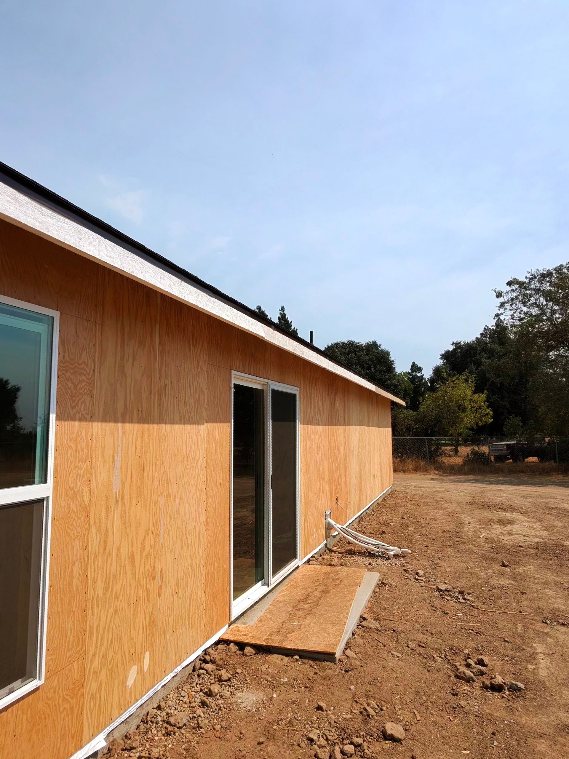 Long wooden building with sliding glass doors beside a bare dirt yard under a clear sky
