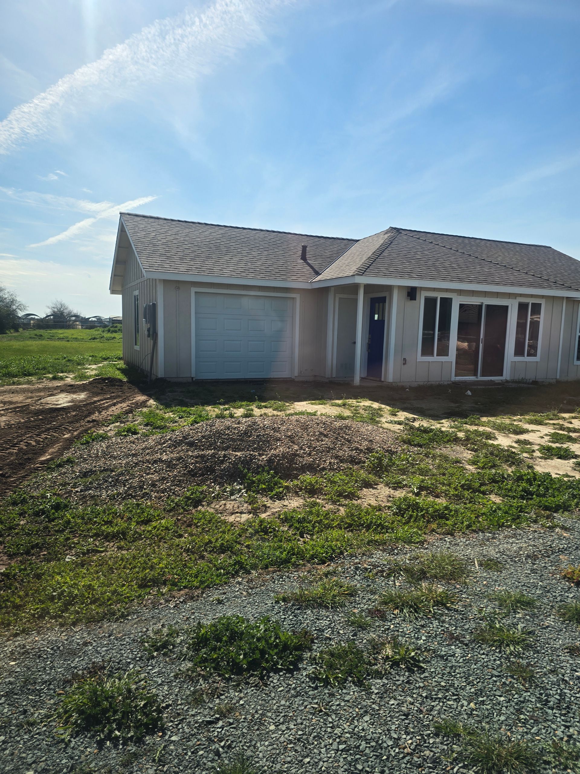 Single-story house with a gray roof and garage, set on a gravel lot under a blue sky.