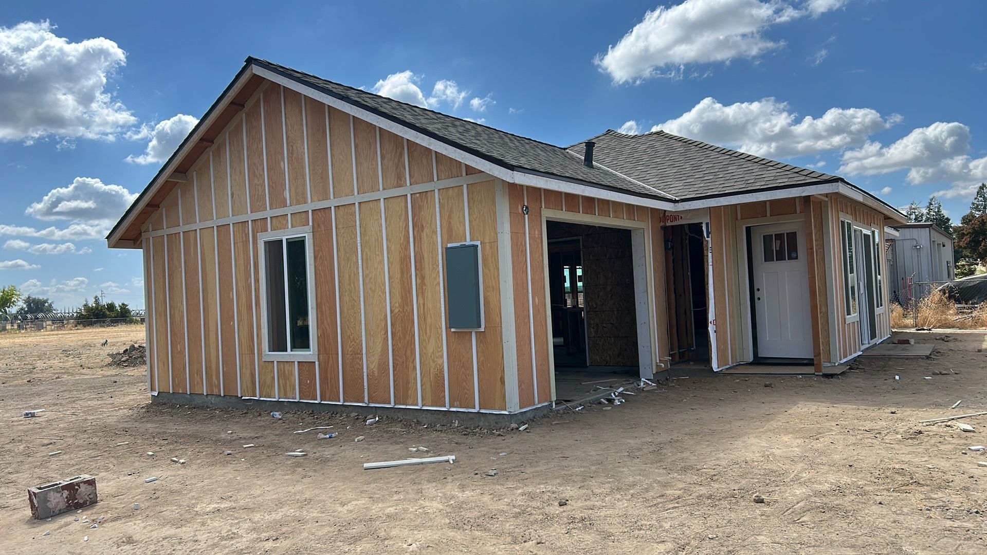 Partially framed wooden house under construction on a dusty lot beneath a cloudy blue sky