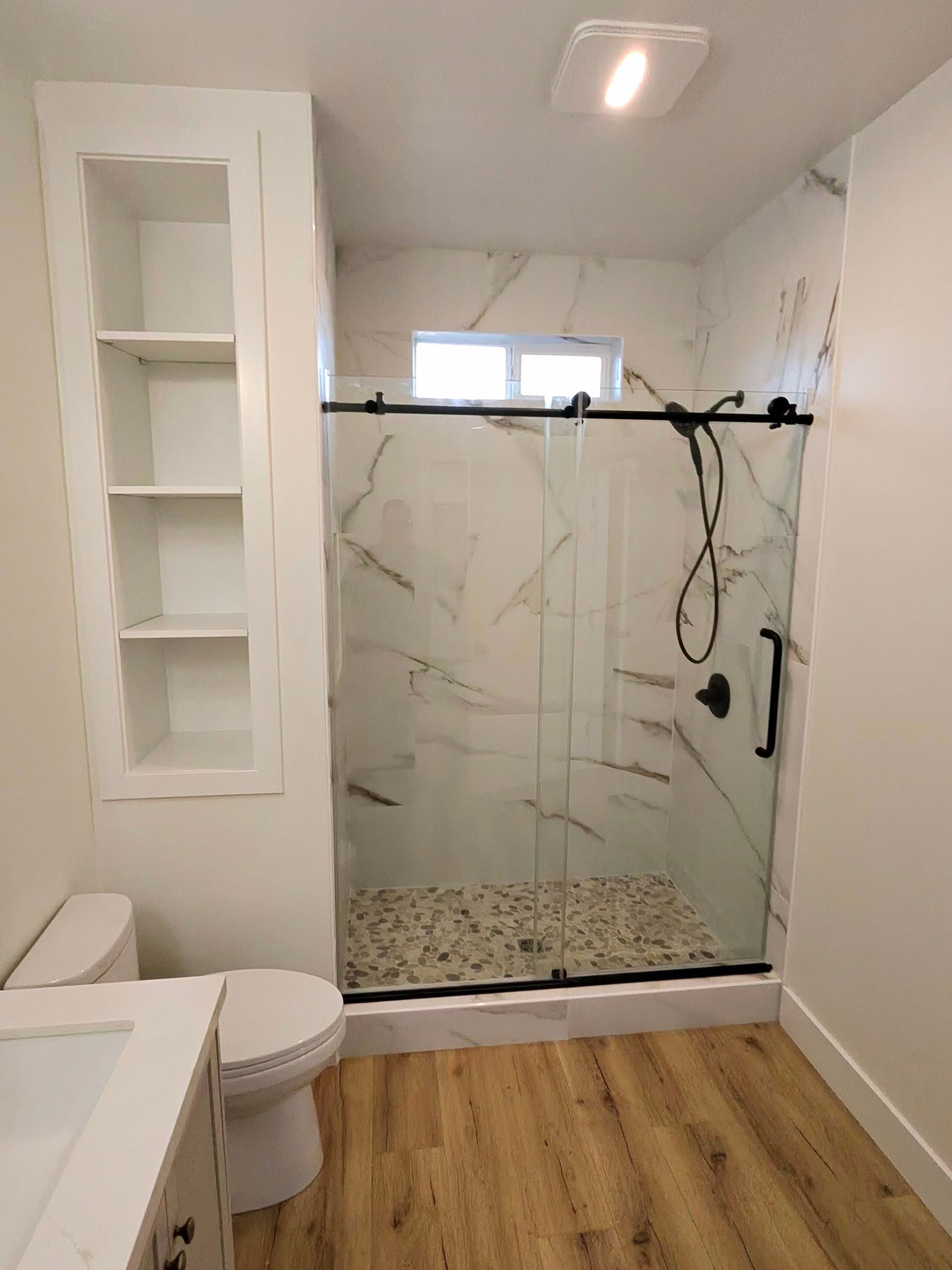 Small bathroom with white shower, toilet, built-in shelves, and wood-look floor.