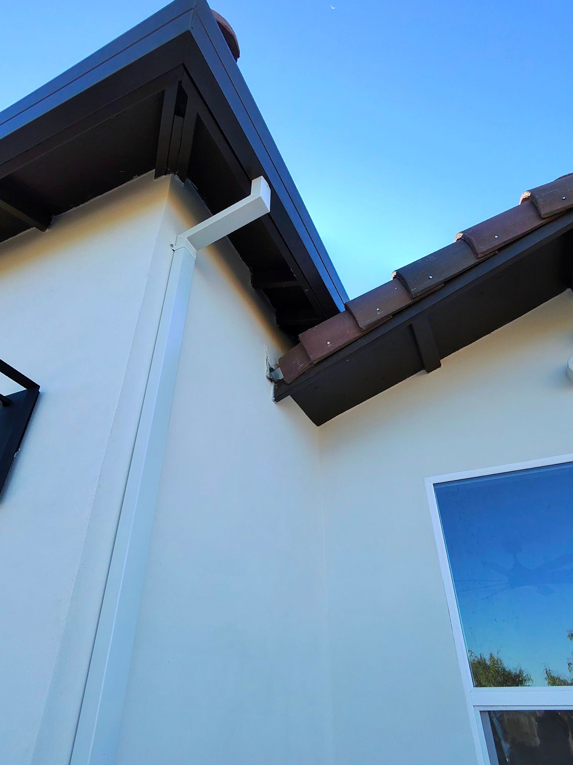 Exterior corner of a white house with dark roof eaves and blue sky reflected in a window