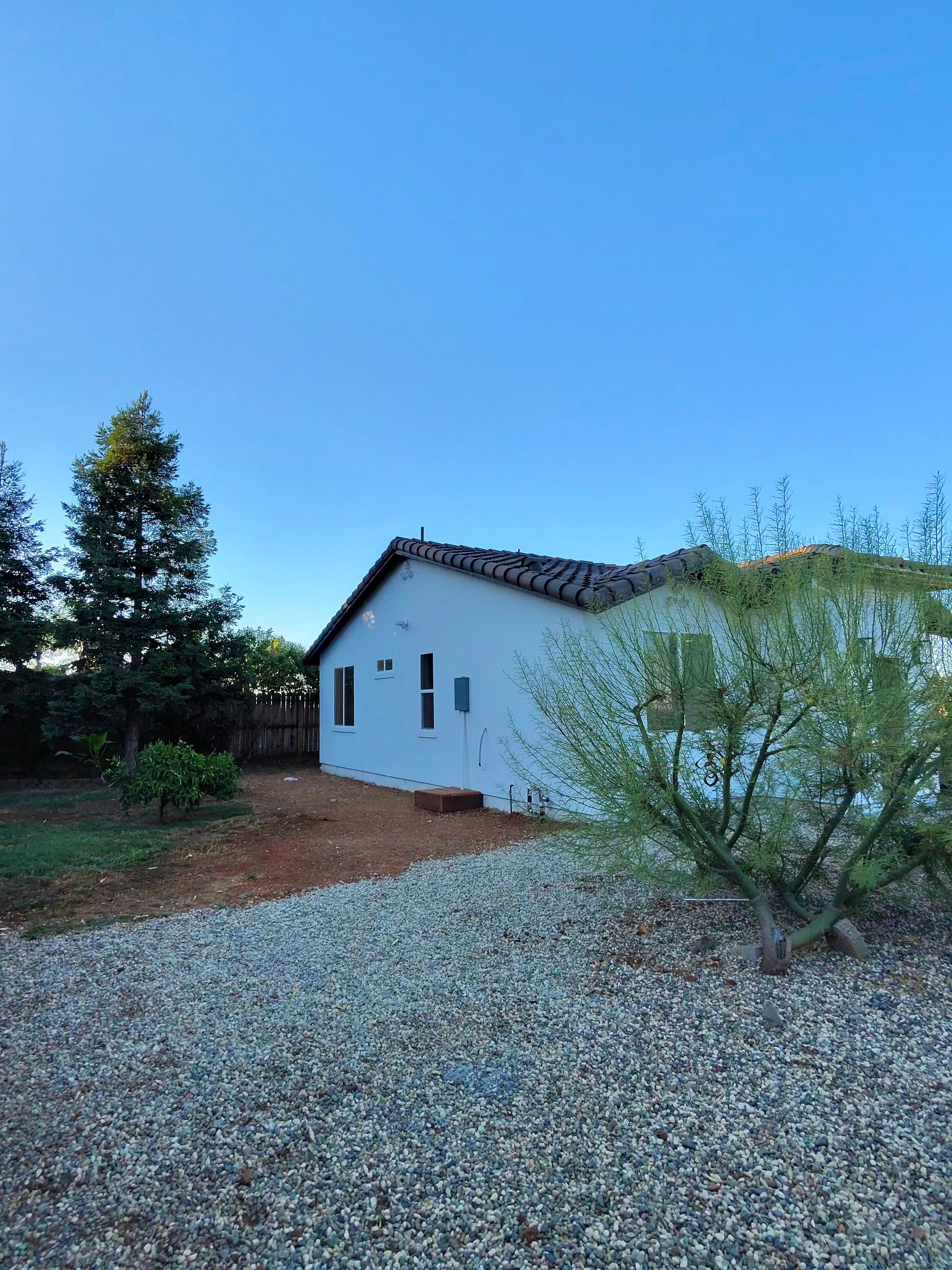 White house with blue shutters beside gravel driveway, trees, and clear blue sky.