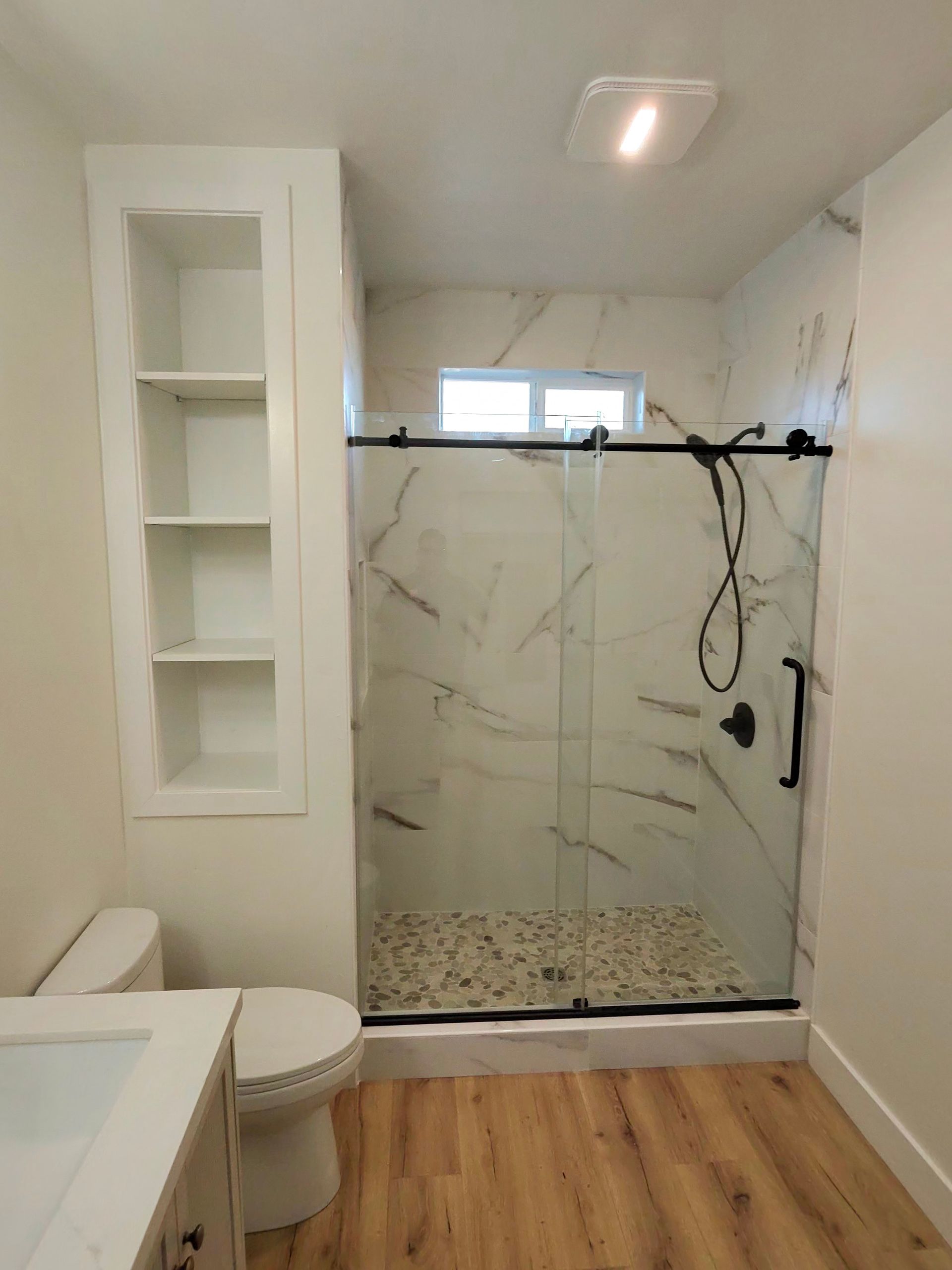 Small bathroom with glass shower, toilet, sink, white tile walls, and wood-look floor
