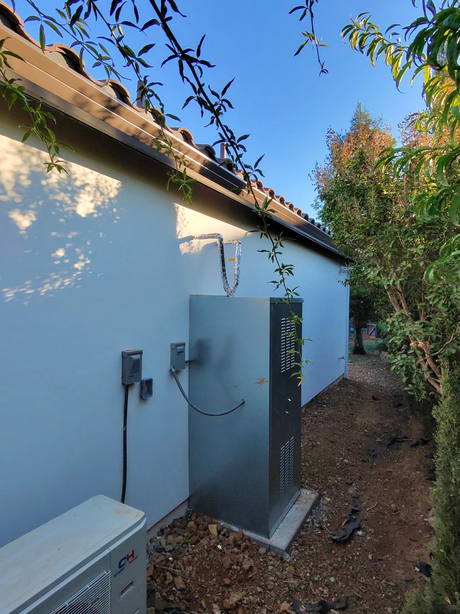 Exterior side of a white house with utility boxes and outdoor HVAC units beside a narrow dirt path
