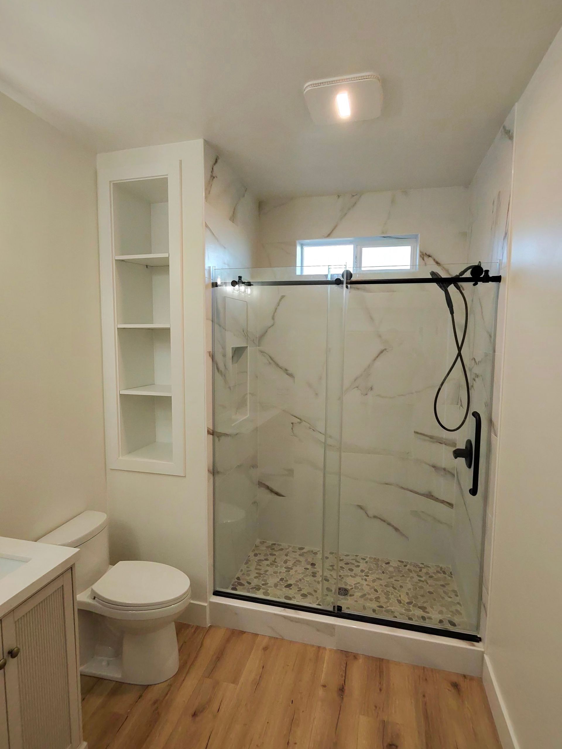 Bathroom with glass walk-in shower, toilet, white shelving, and wood-look floor.
