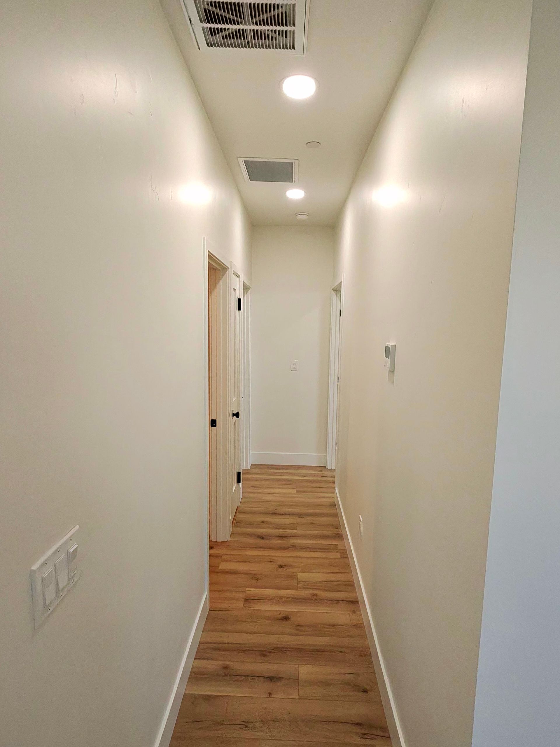 Narrow white hallway with wood floors and recessed ceiling lights, viewed from one end.