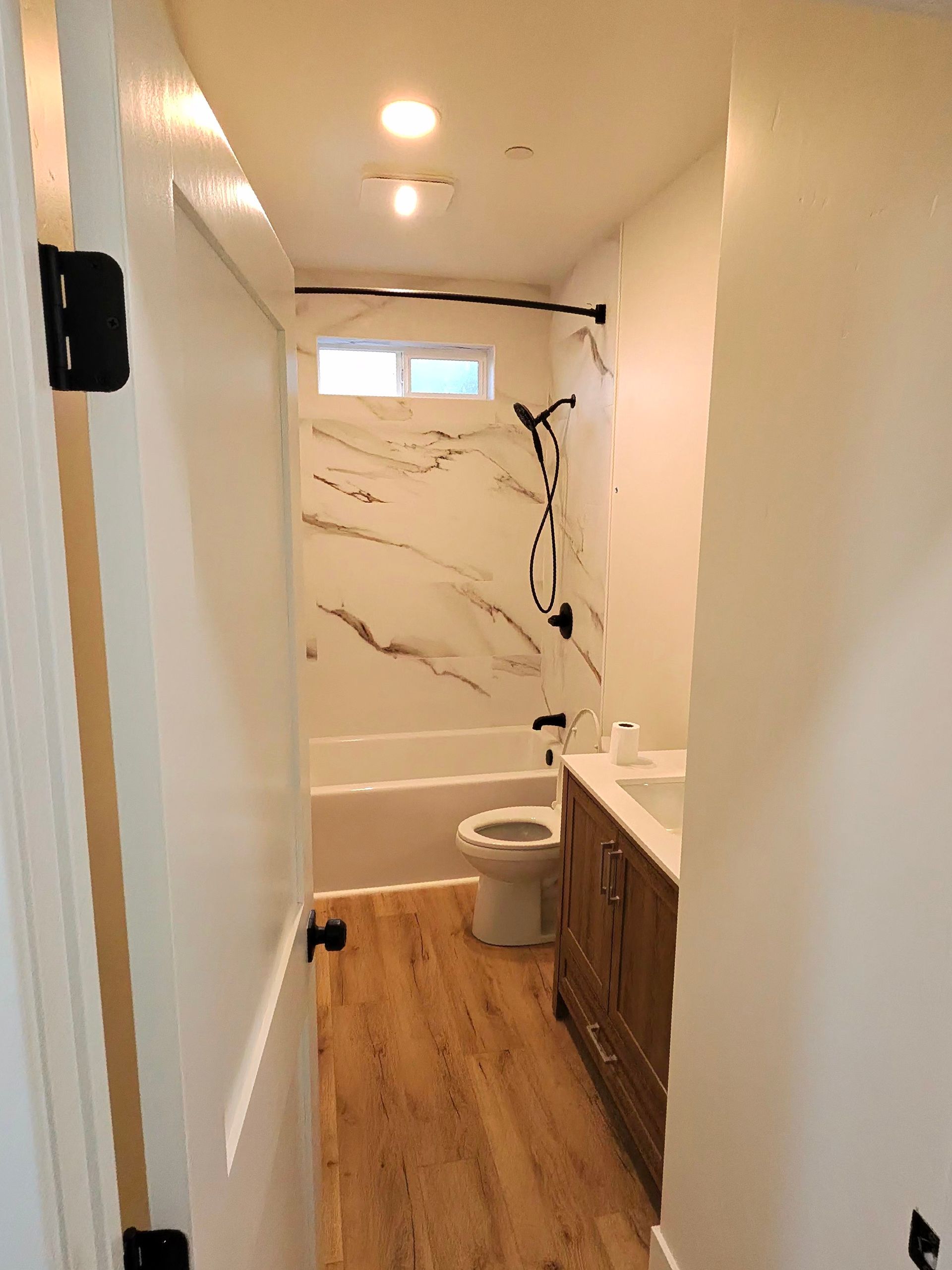 Hallway view into a small bathroom with wood vanity, marble shower wall, and toilet