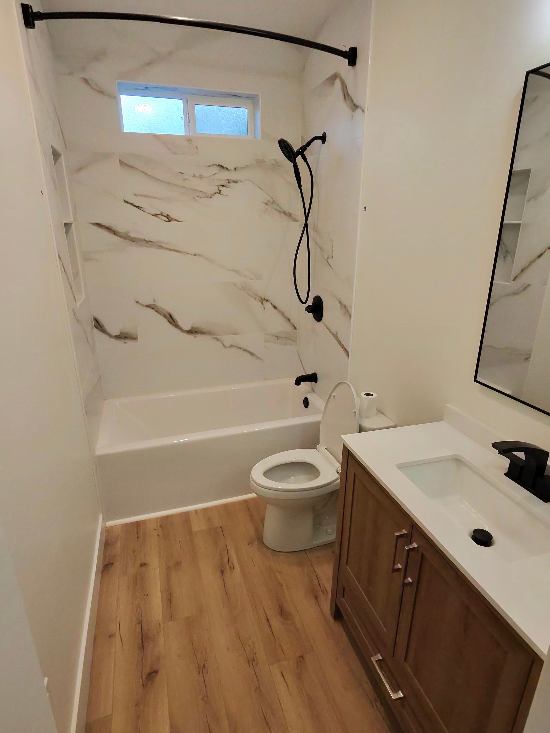 Small bathroom with shower, toilet, vanity, white walls, and wood-look floor.