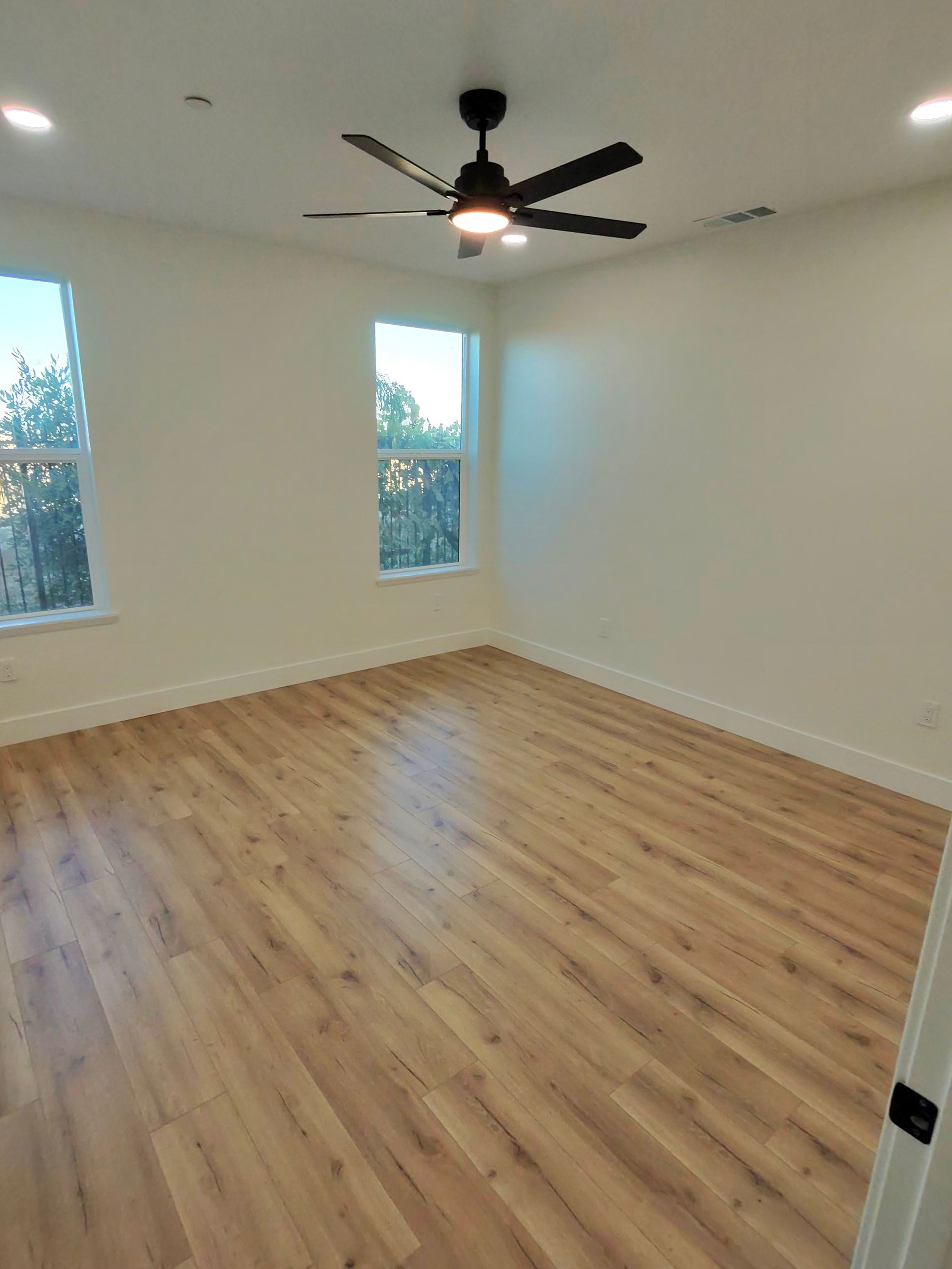 Empty unfurnished room with wood floors, white walls, two windows, and a ceiling fan.