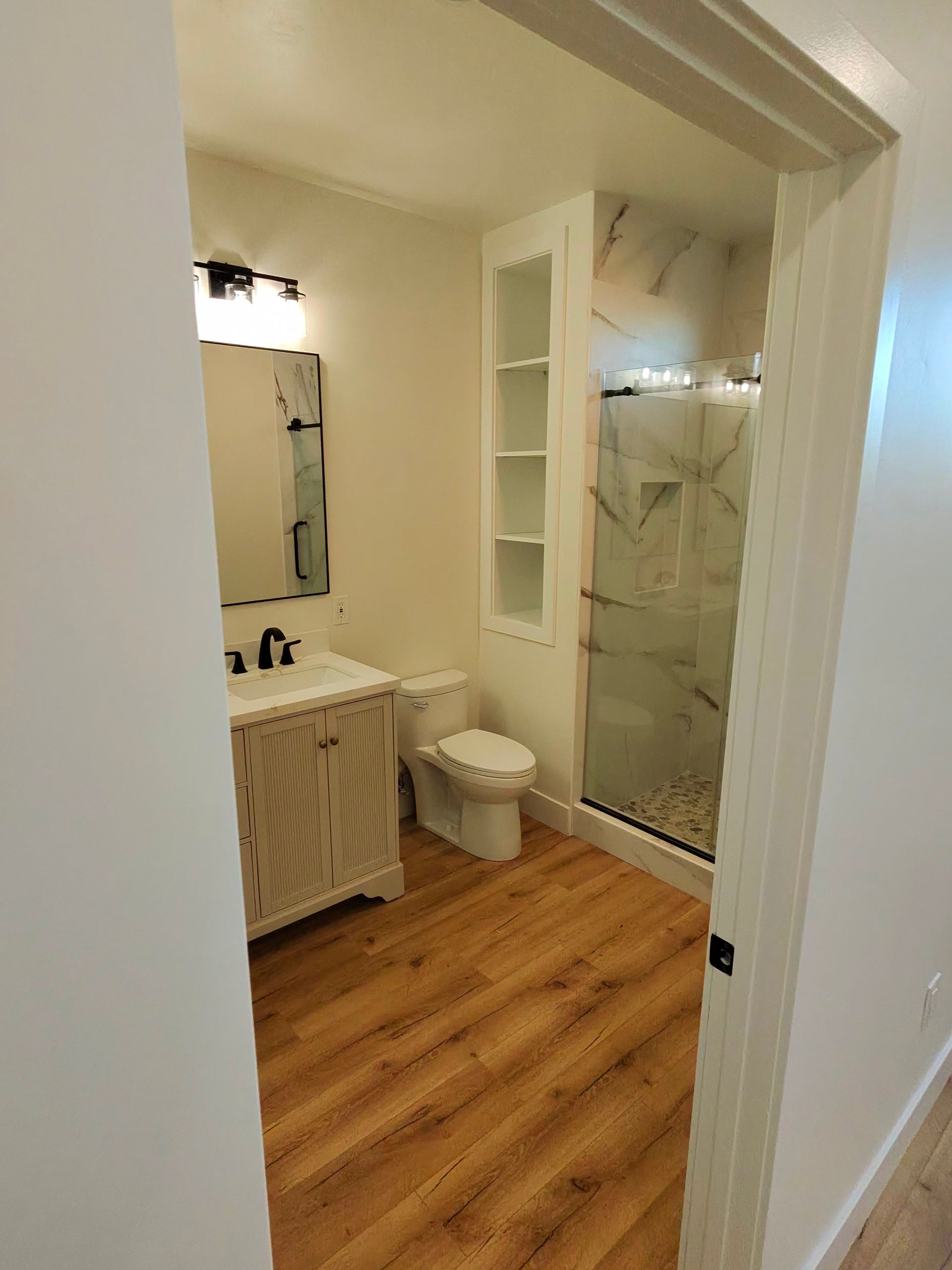 Bathroom with white vanity, toilet, glass shower, built-in shelves, and wood-look flooring.