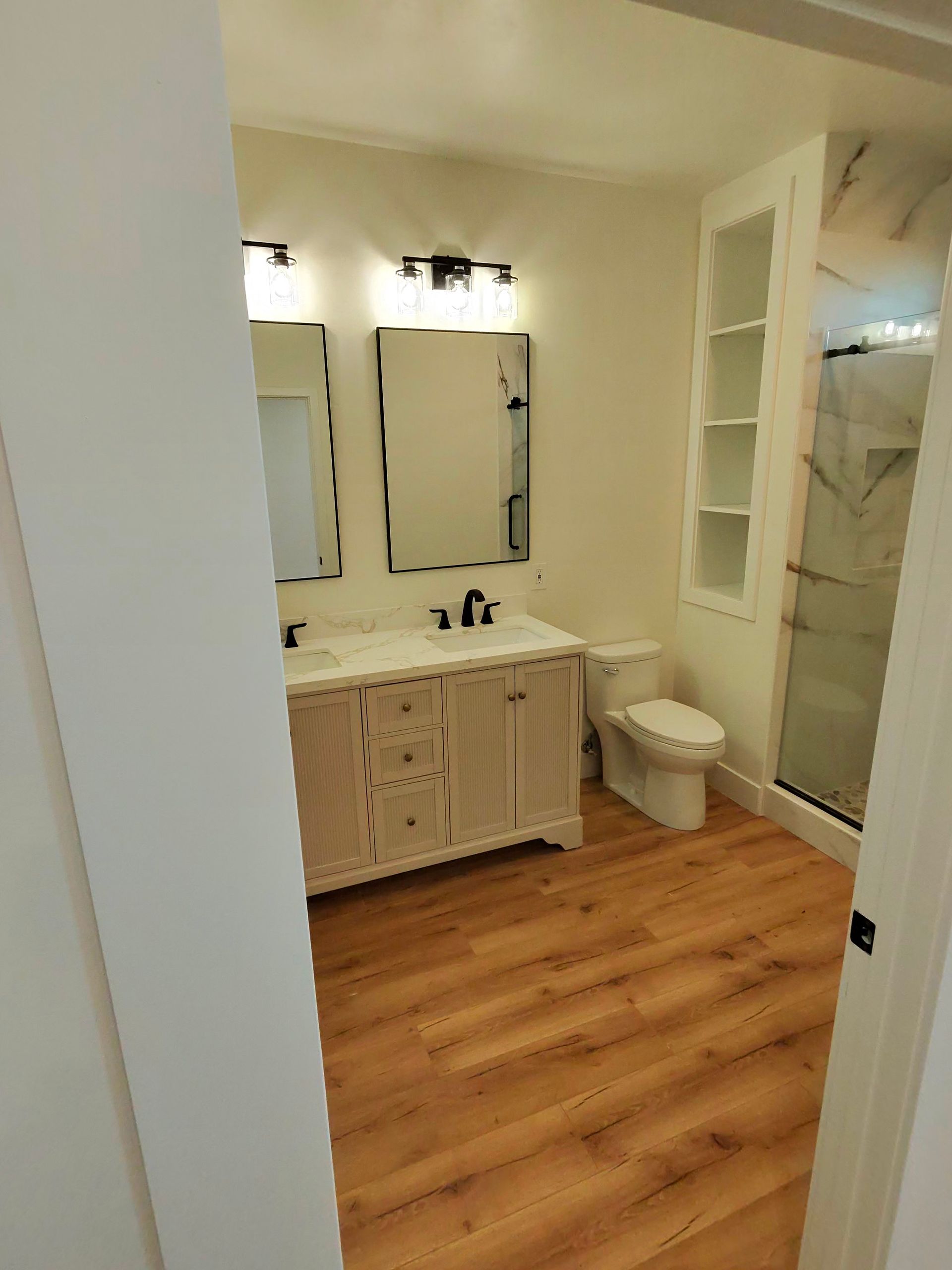 Bathroom with light wood floor, white vanity and mirror, toilet, and glass shower enclosure.