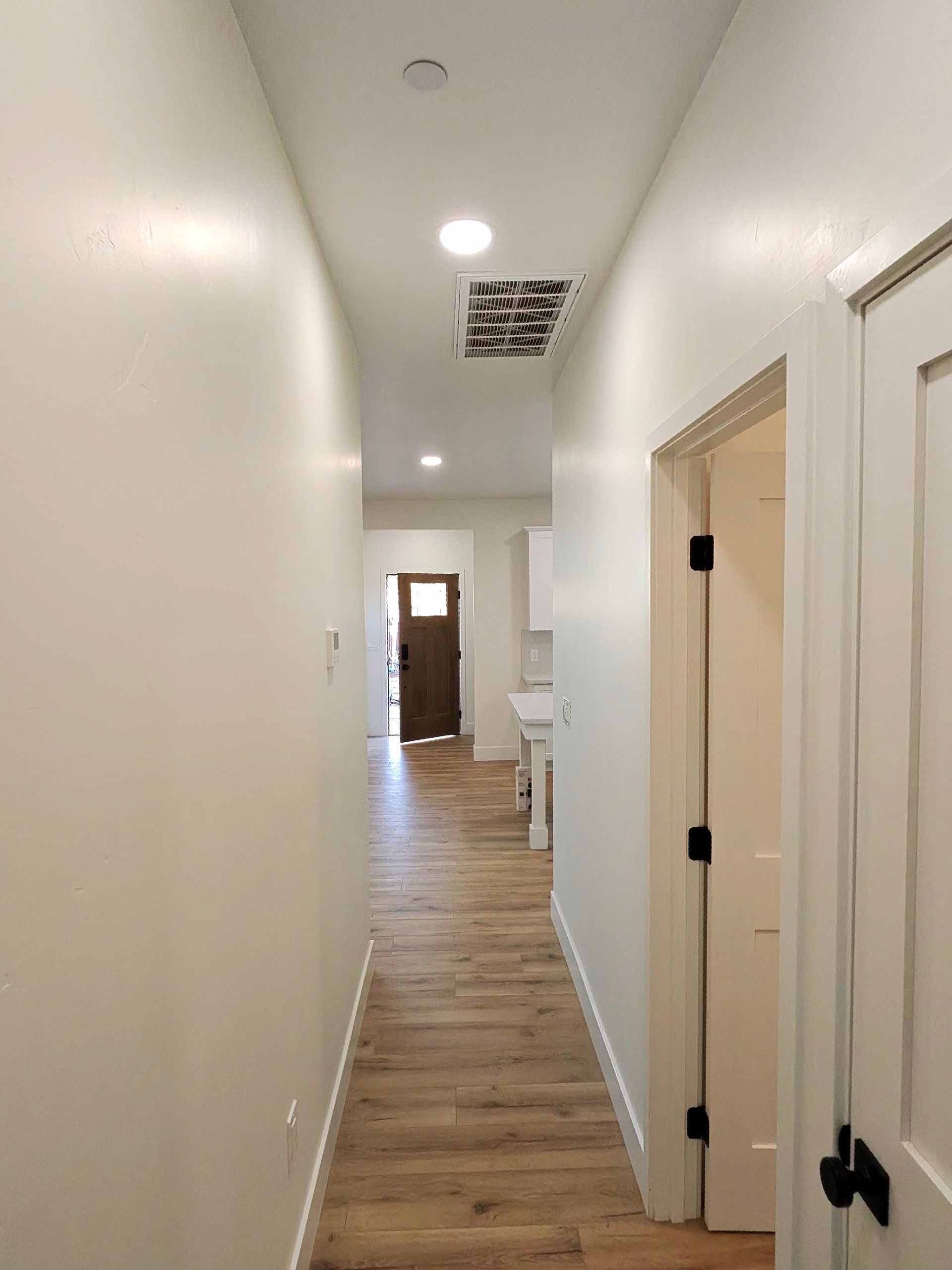 Narrow white hallway with wood floors, ceiling lights, and an open doorway at the end
