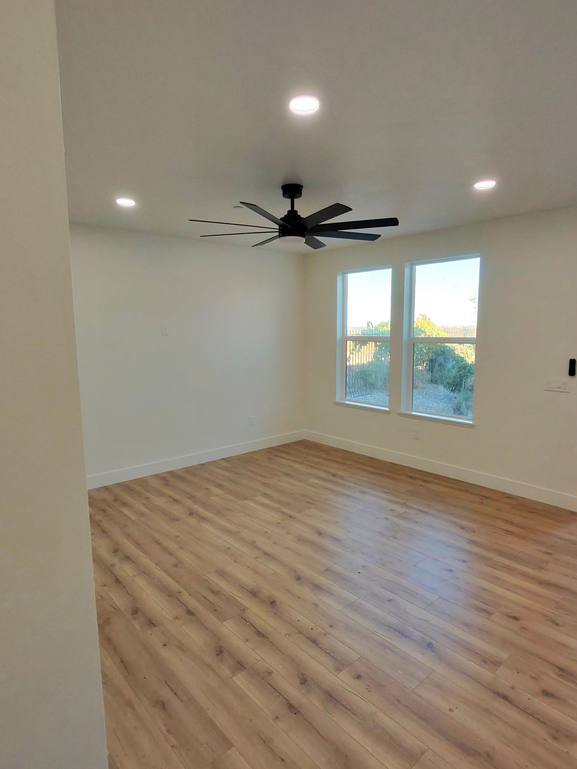 Empty room with light wood floors, white walls, ceiling fan, recessed lights, and a window overlooking greenery.