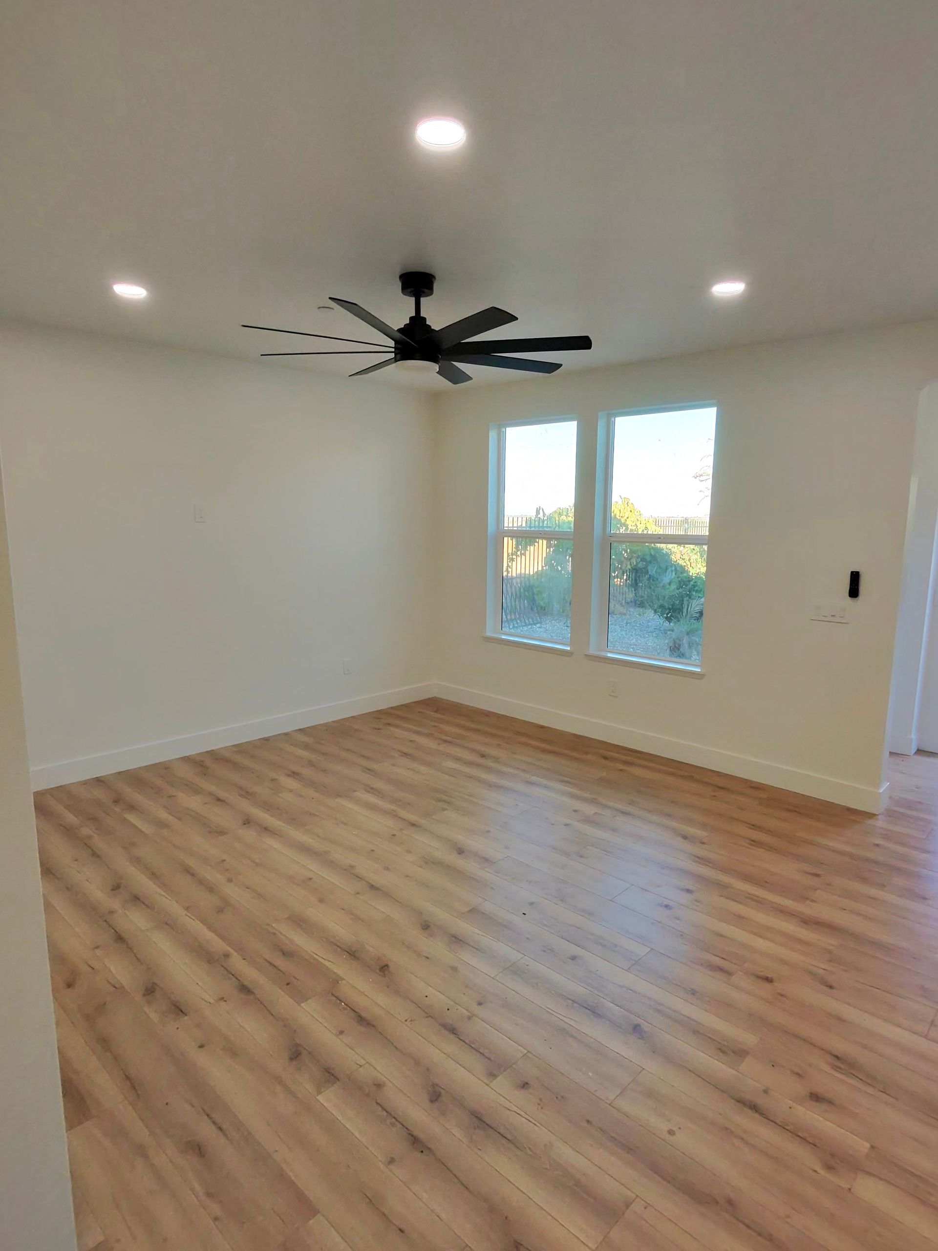 Empty room with light wood floor, white walls, recessed lights, ceiling fan, and two windows.