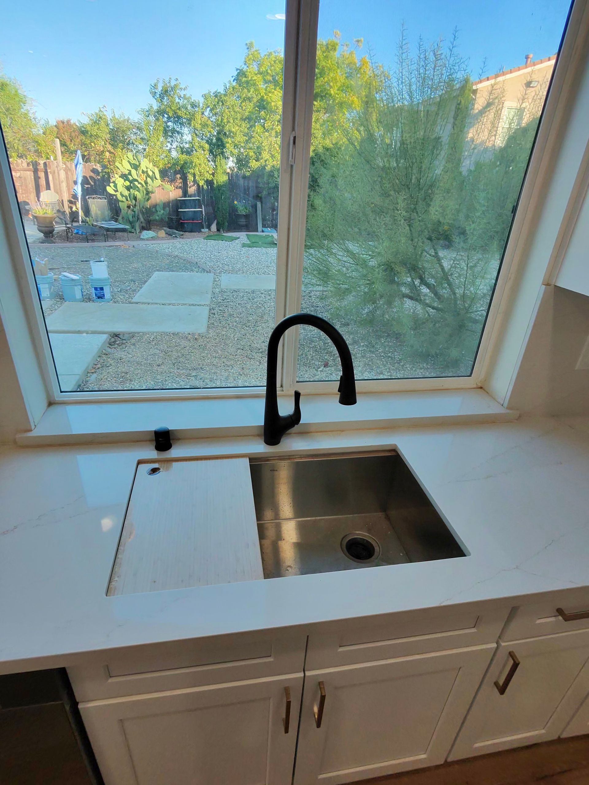 Kitchen sink and white cabinets beneath a large window overlooking a sunny backyard.