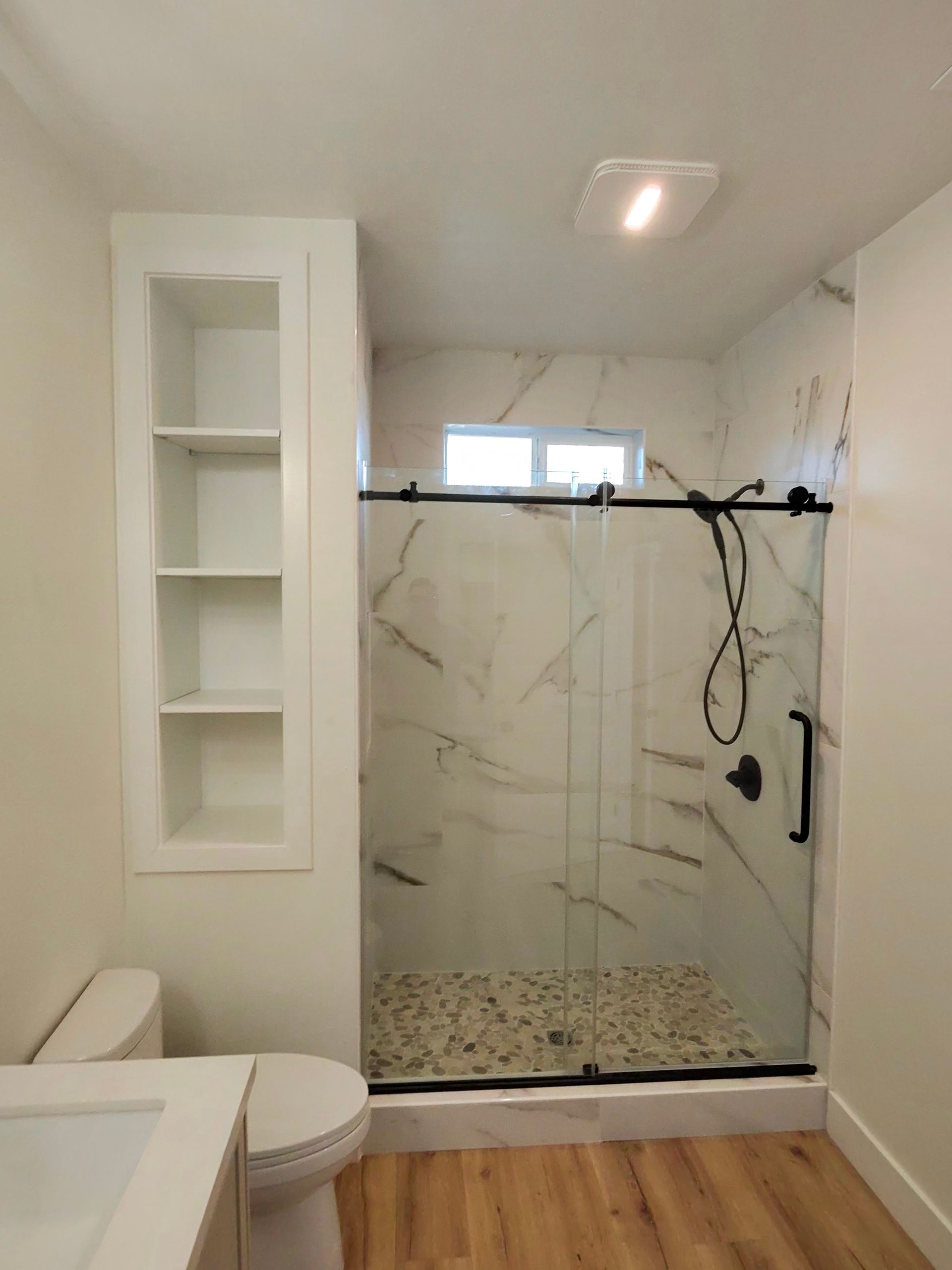 Modern bathroom with glass shower, marble-look tile, white shelving, and a toilet beside wood flooring