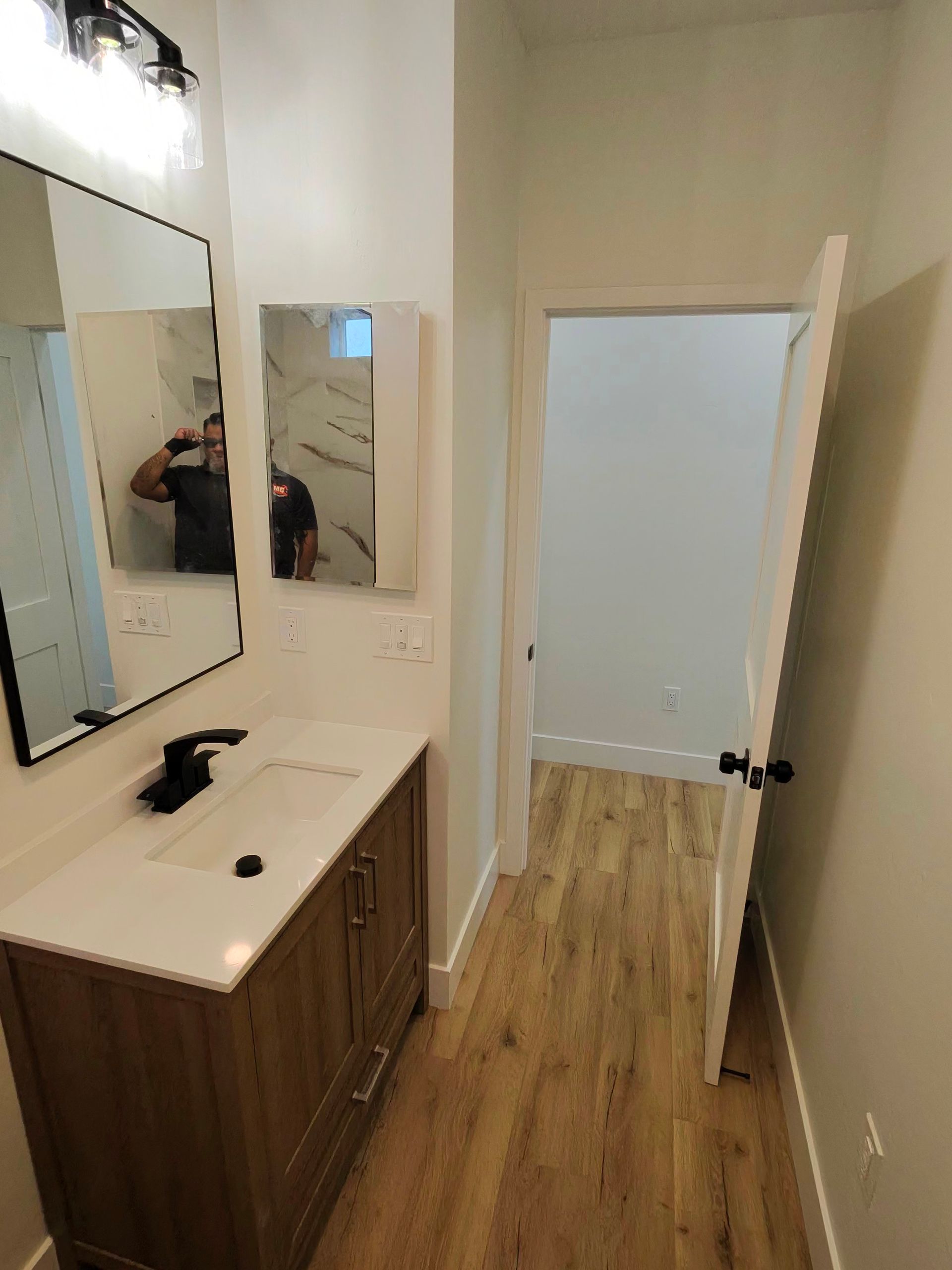 Narrow hallway with wood floor, white walls, and a vanity sink with large mirrors on the left.