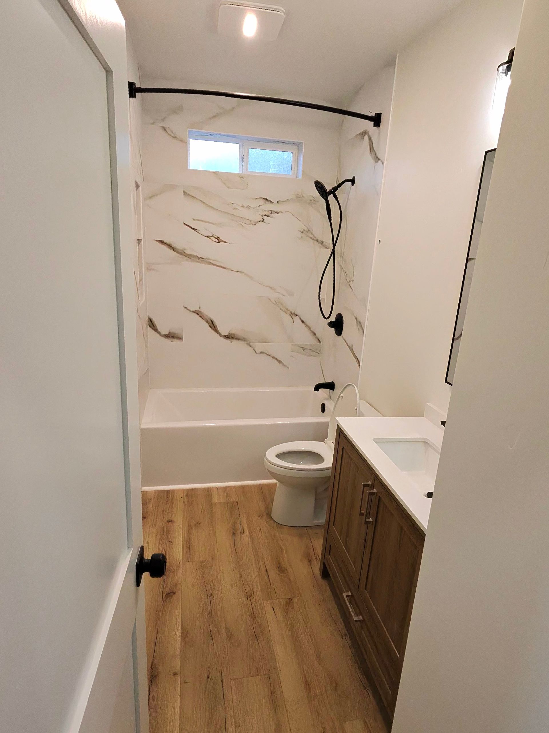 Small bathroom with wood-look floor, white tub, toilet, and vanity with marble-pattern walls.