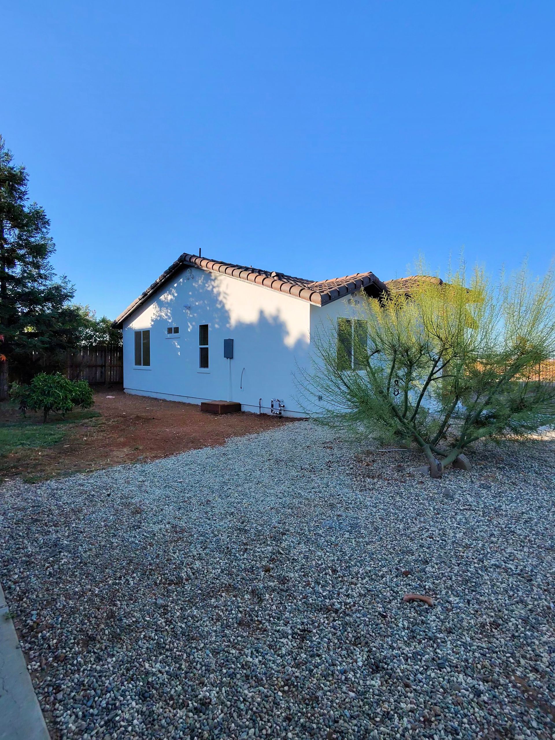 White stucco house with a tiled roof beside a gravel yard and trees under a blue sky