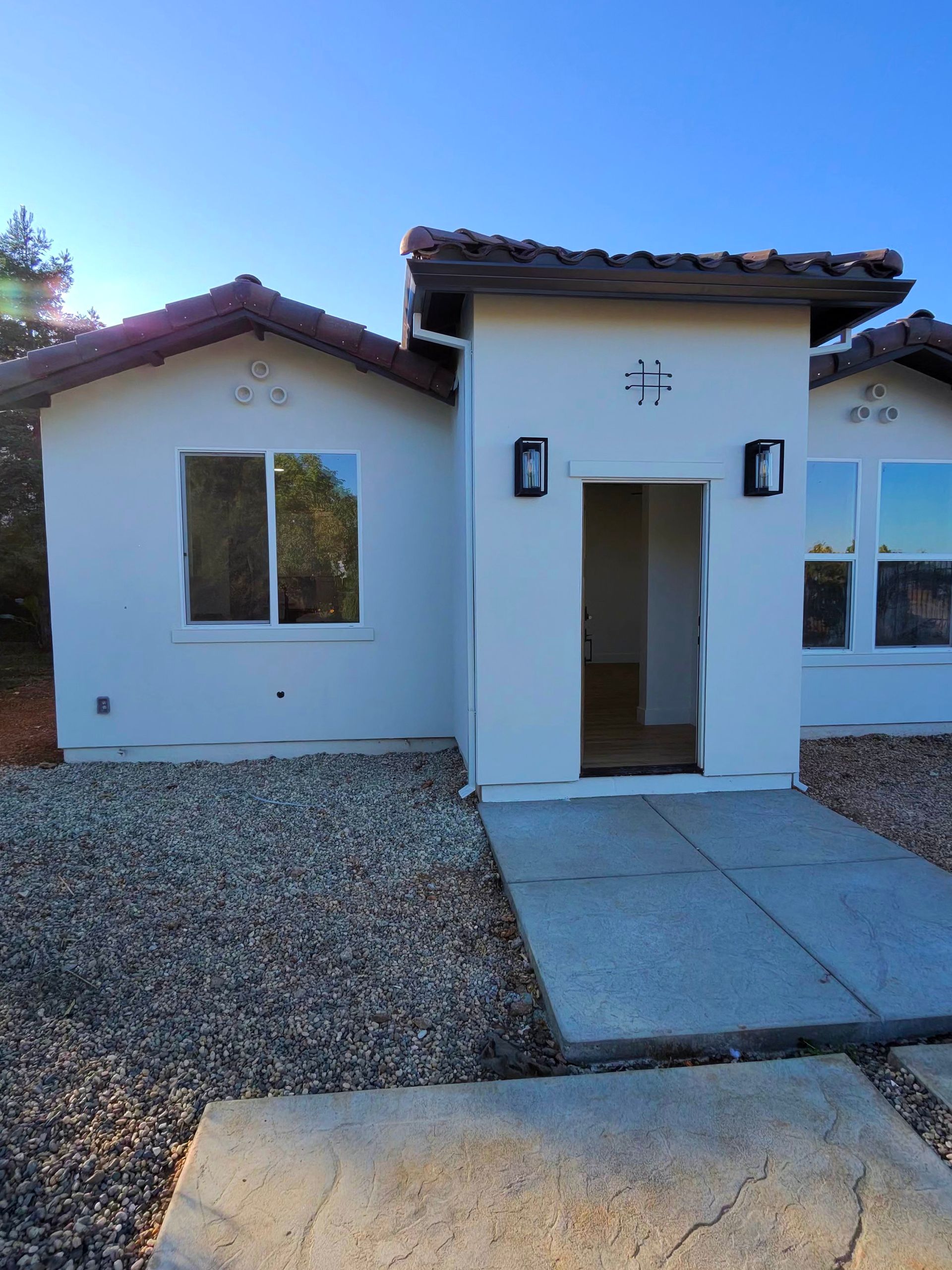 Small white stucco house with a tiled roof, front door, windows, and a concrete walkway over gravel.