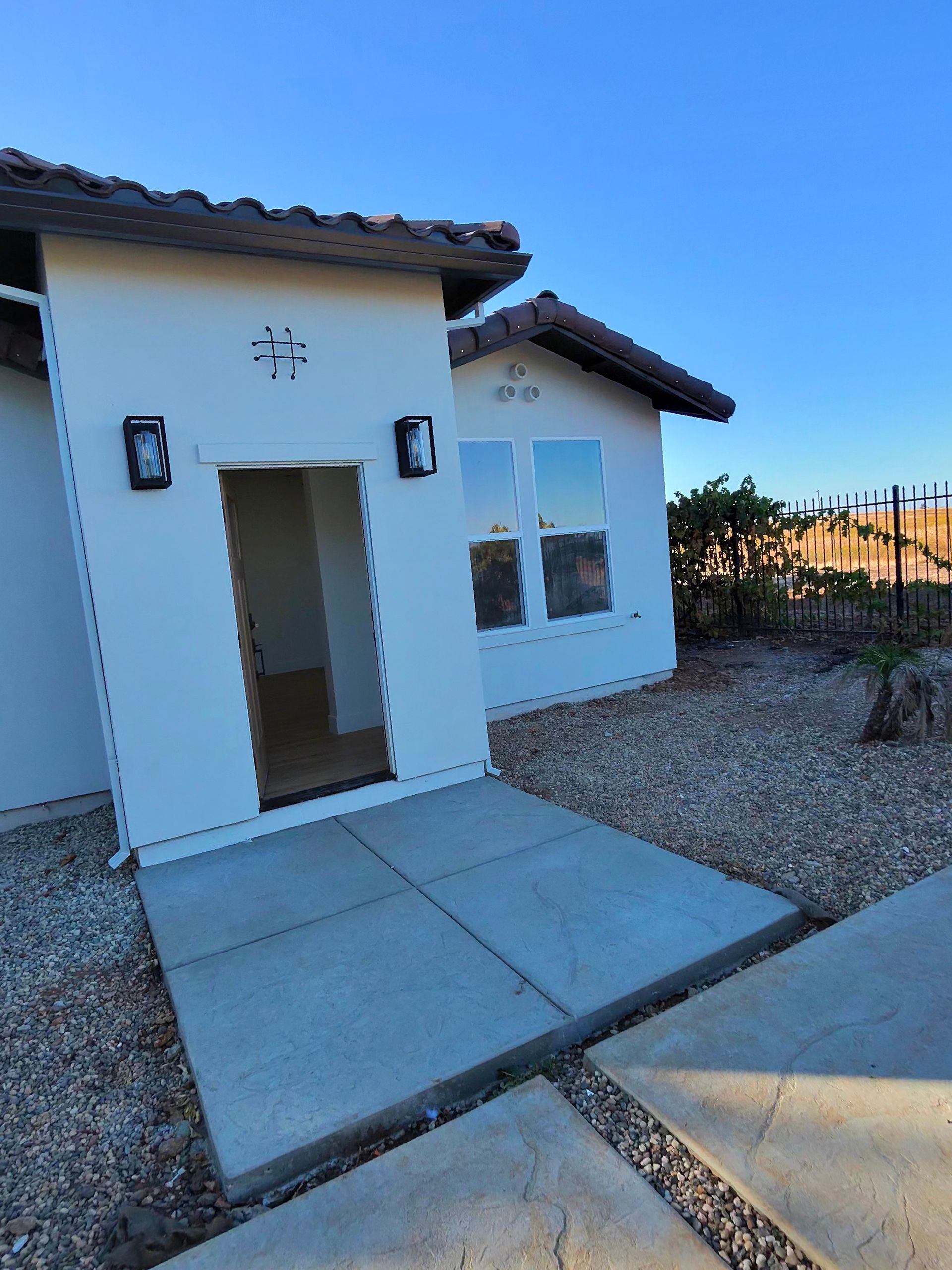 Small white stucco house entrance with a walkway, gravel yard, and sunset sky.