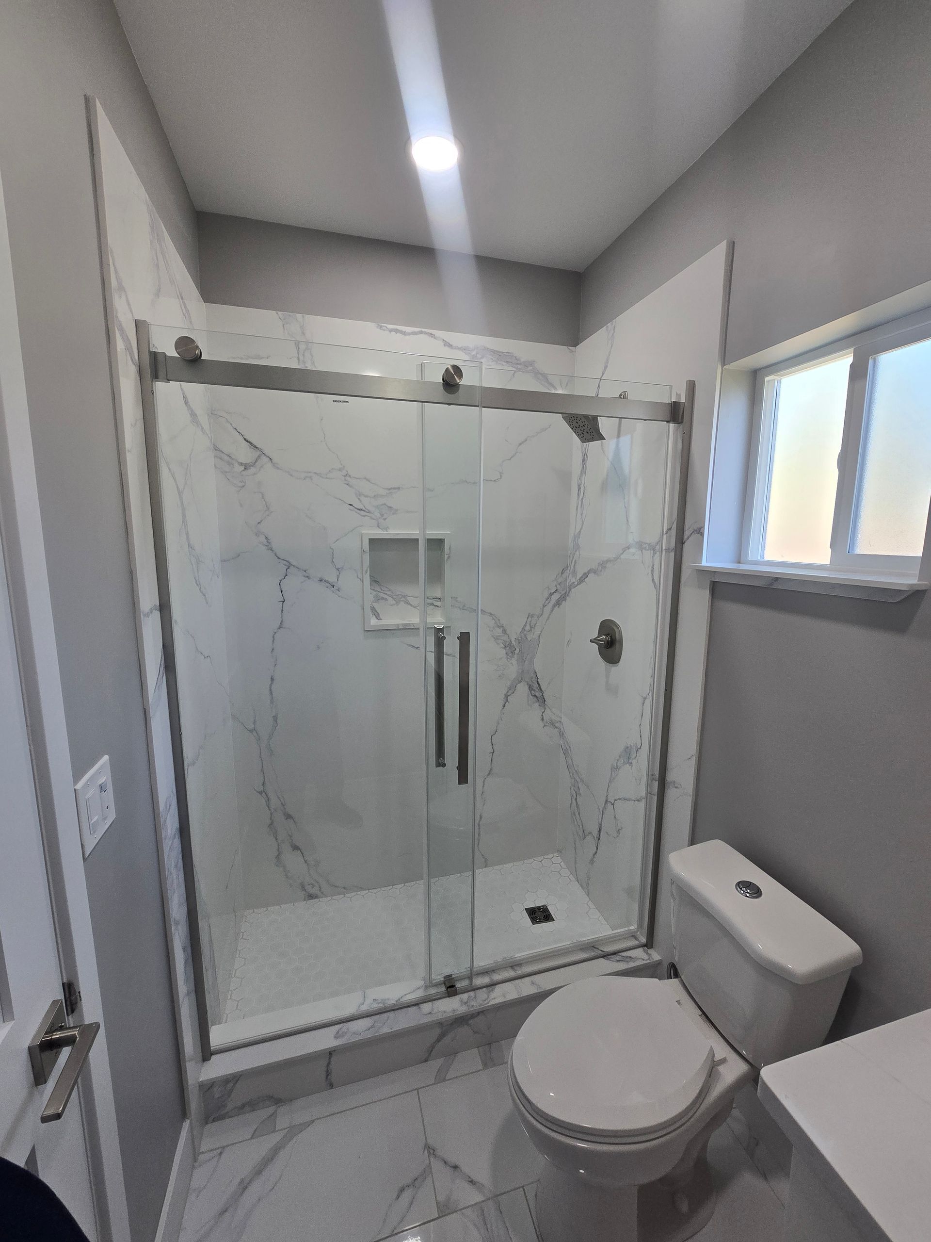 Modern gray bathroom with glass shower, toilet, vanity sink, and small window