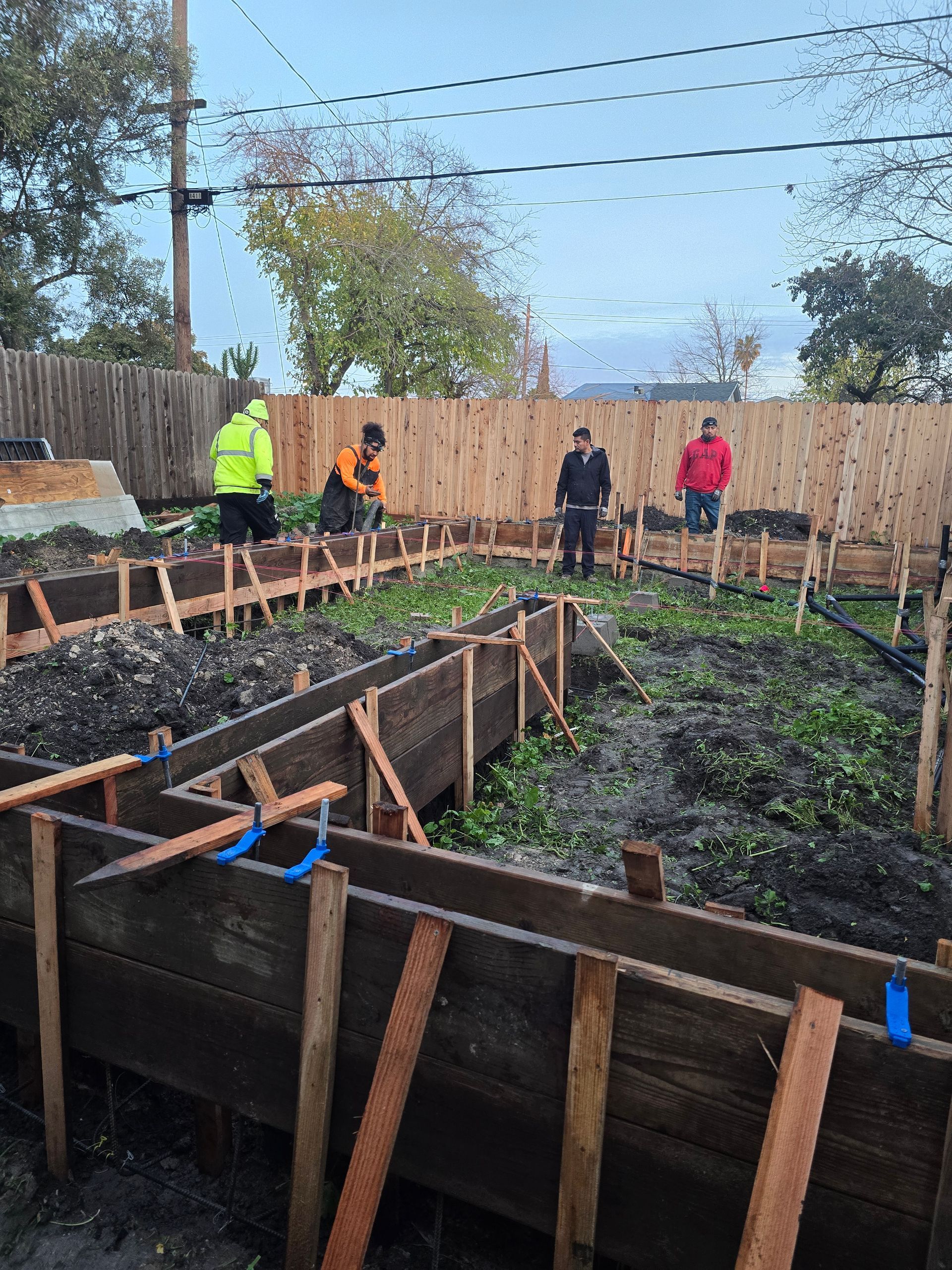 Garden worksite with raised beds, soil, and four people standing by a wooden fence under string lights
