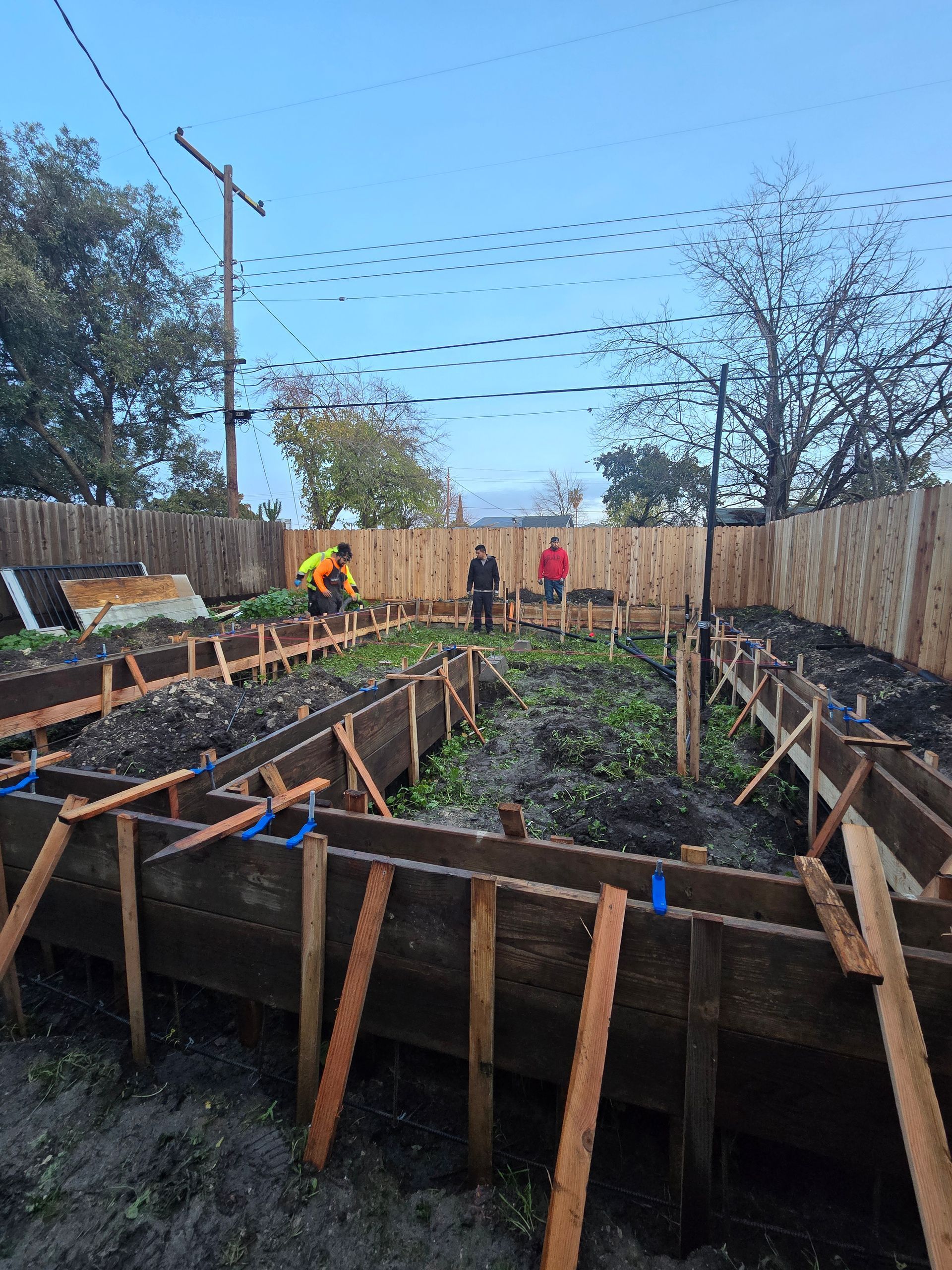 Garden beds in a backyard, with people working near a wooden fence under strings overhead.