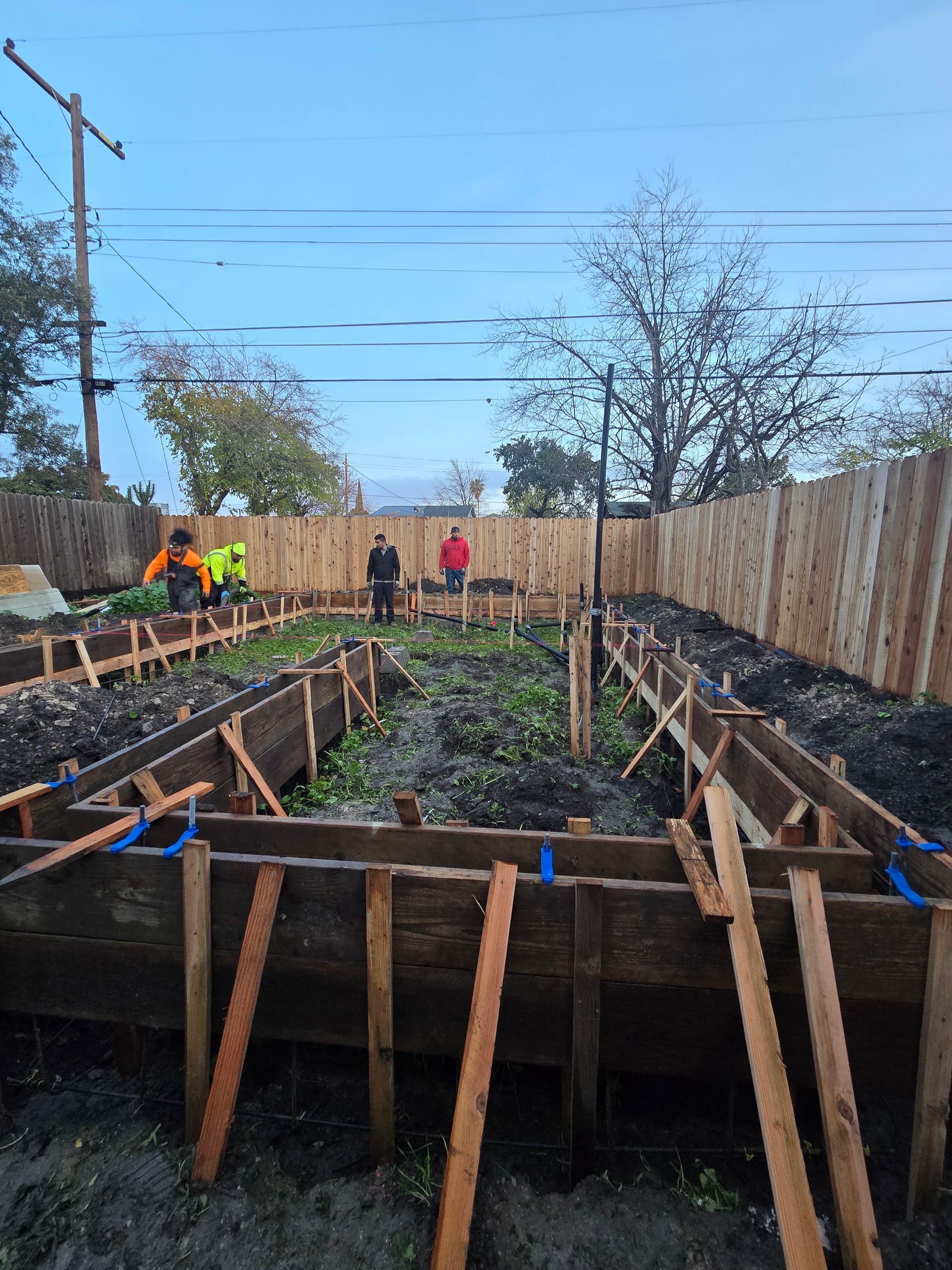 Backyard construction site with wooden formwork and workers in high-visibility clothing.