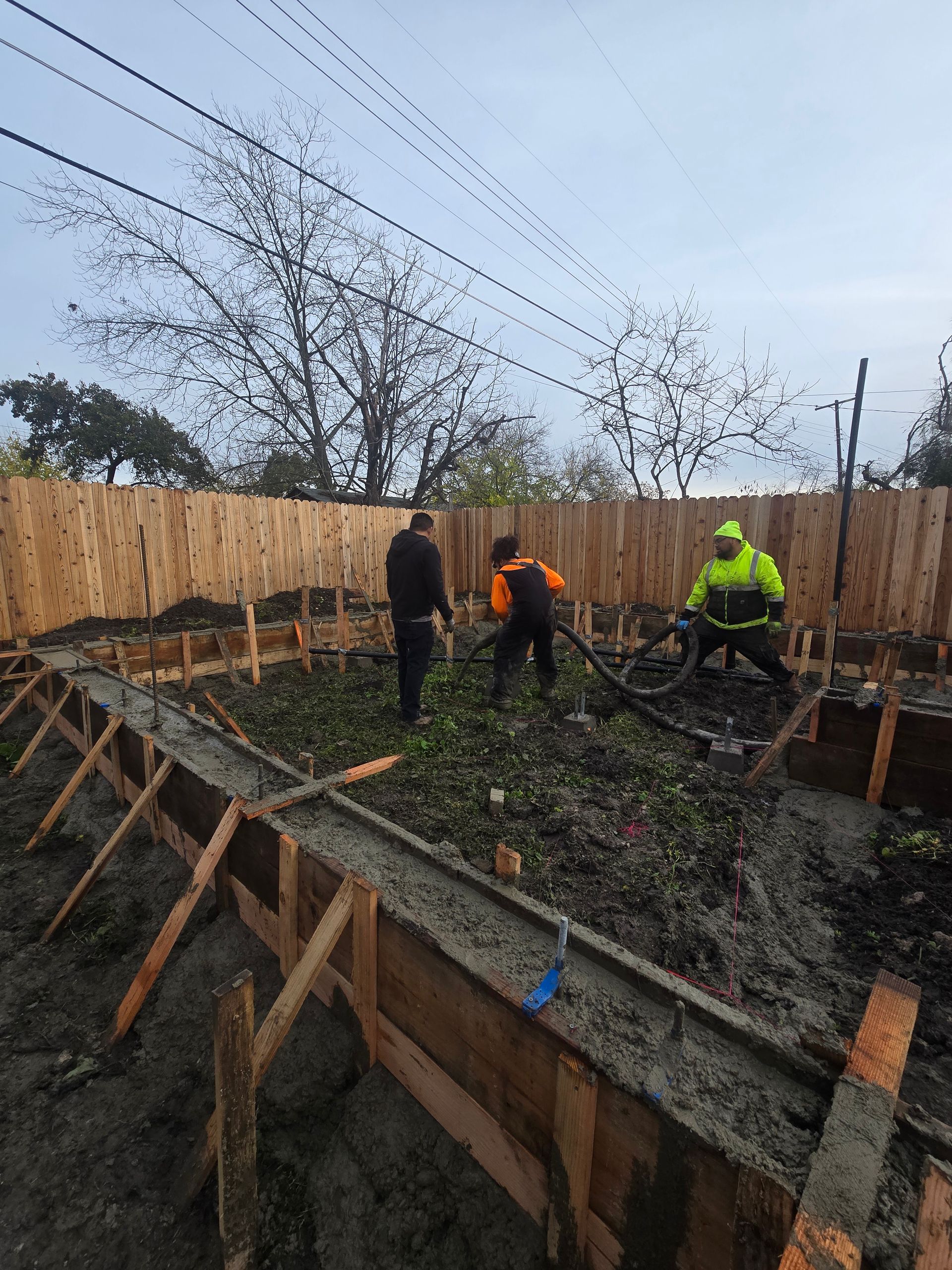 Workers in orange and neon vests pouring concrete into a wooden foundation form at a construction site