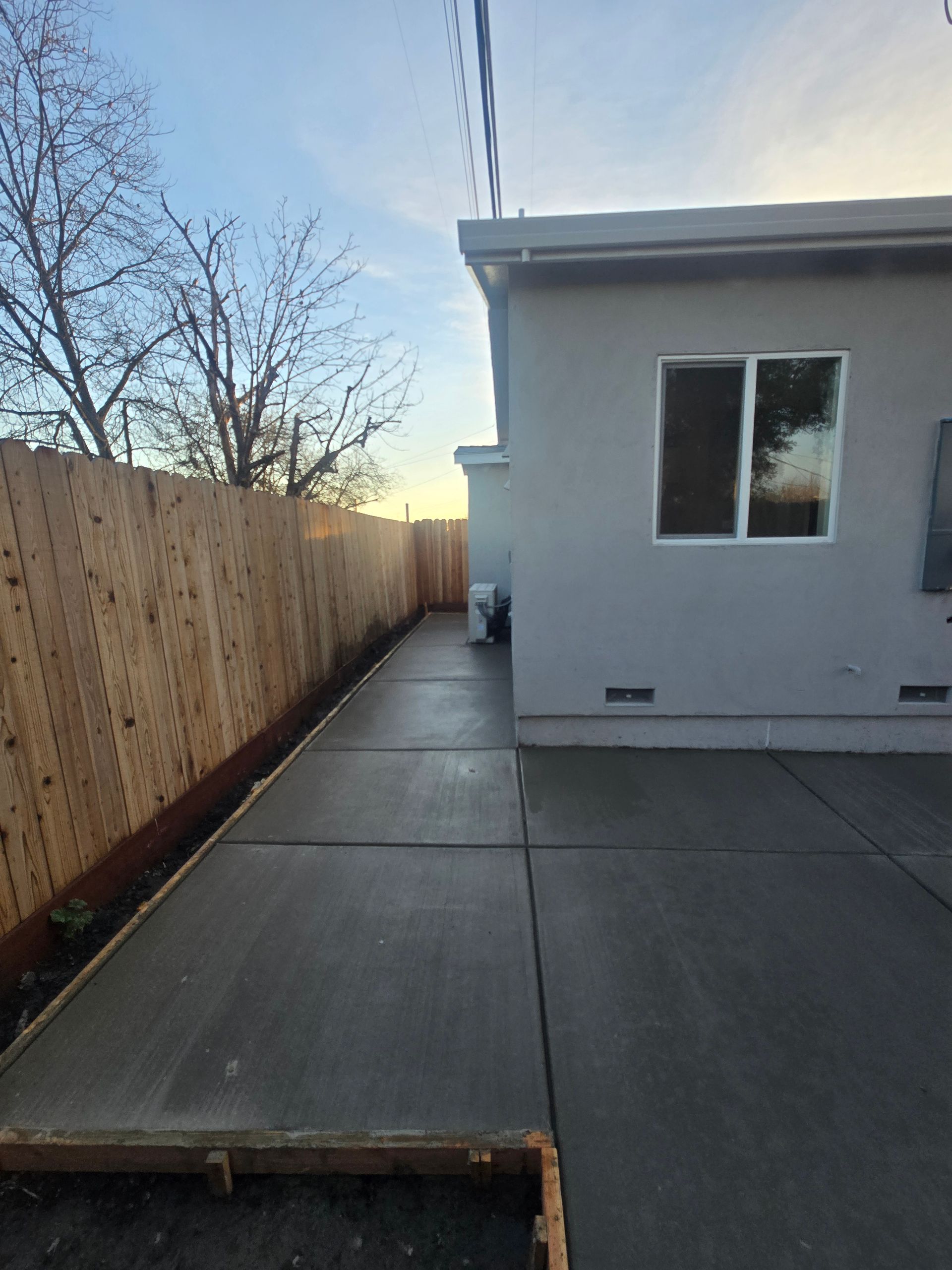 Concrete side yard walkway beside a house, with a wooden fence on the left and a window on the right.