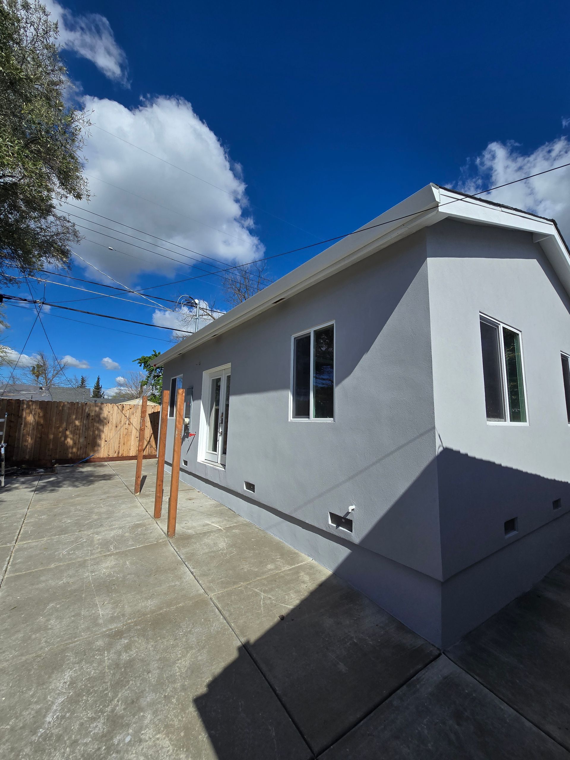 Single-story gray stucco house with white trim under a bright blue sky, viewed from the side.