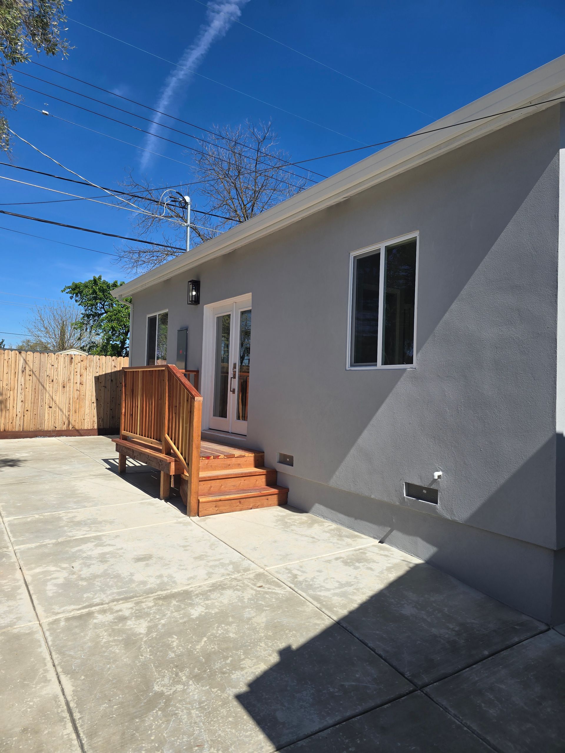 Back patio with concrete slab, gray stucco house, wooden steps, and fenced yard under blue sky