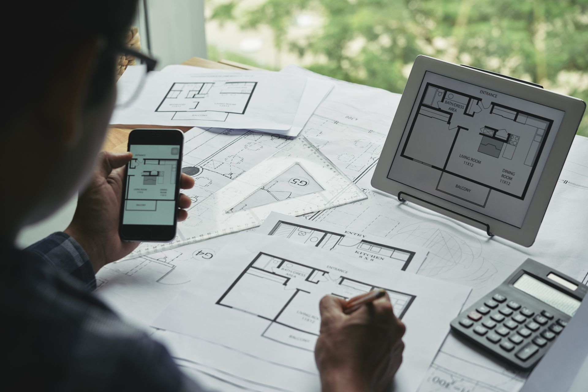 Person reviewing floor plans on phone and tablet at a desk with calculator and paper sketches
