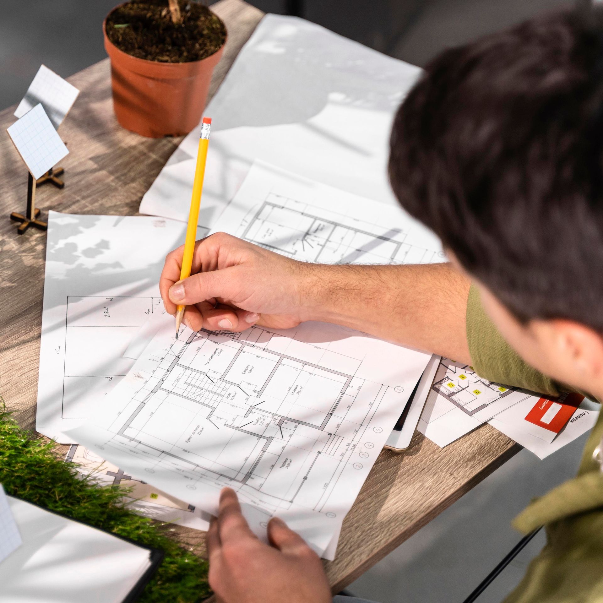 Person sketching a house plan with a pencil at a desk beside a potted plant
