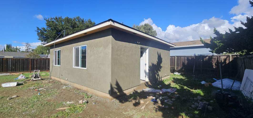 Small beige stucco house with a blue roof in a backyard under a sunny sky.