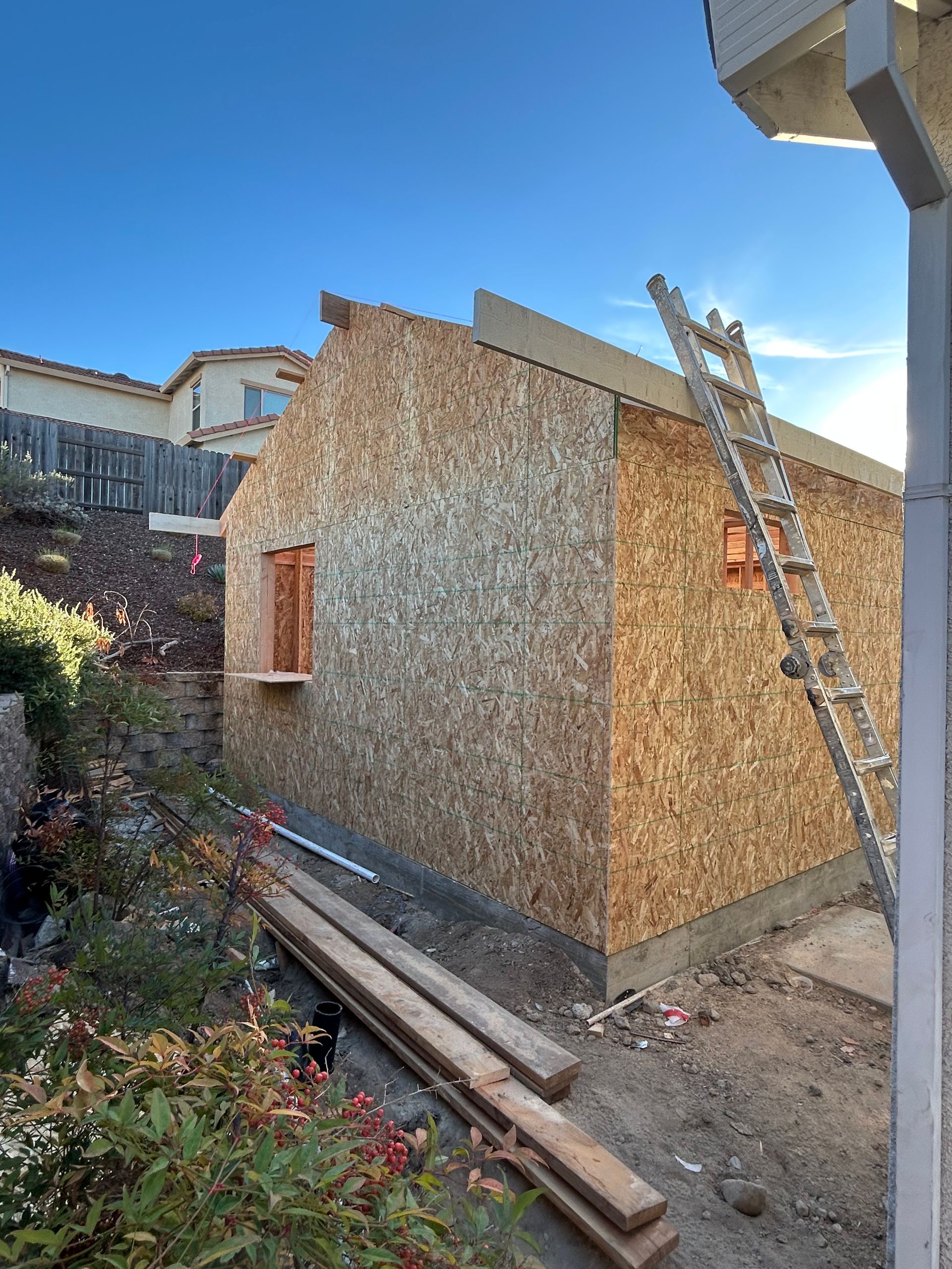 Small wooden shed under construction beside a house, with a ladder leaning against it.