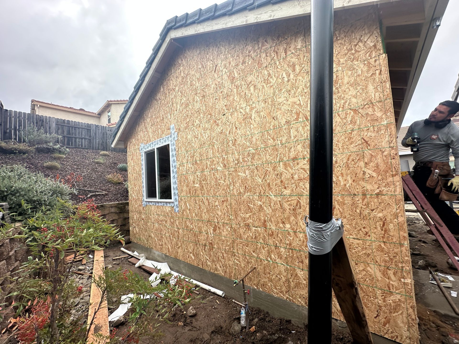 Partially built house with OSB siding and a black drainage pipe beside a muddy slope